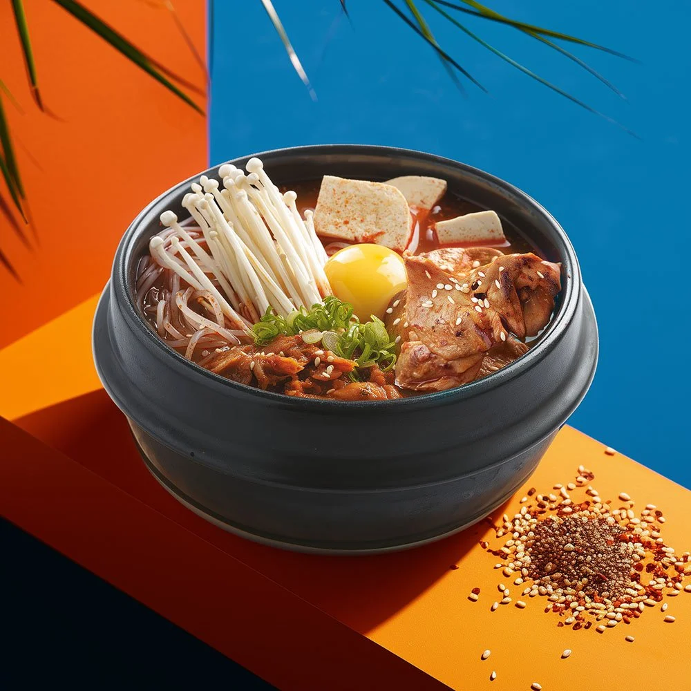 A bowl of Korean-style spicy soup with noodles, bean sprouts, an egg yolk, green onions, tofu, pork, and sesame seeds, placed on an orange and blue background with chili flakes and sesame seeds nearby.