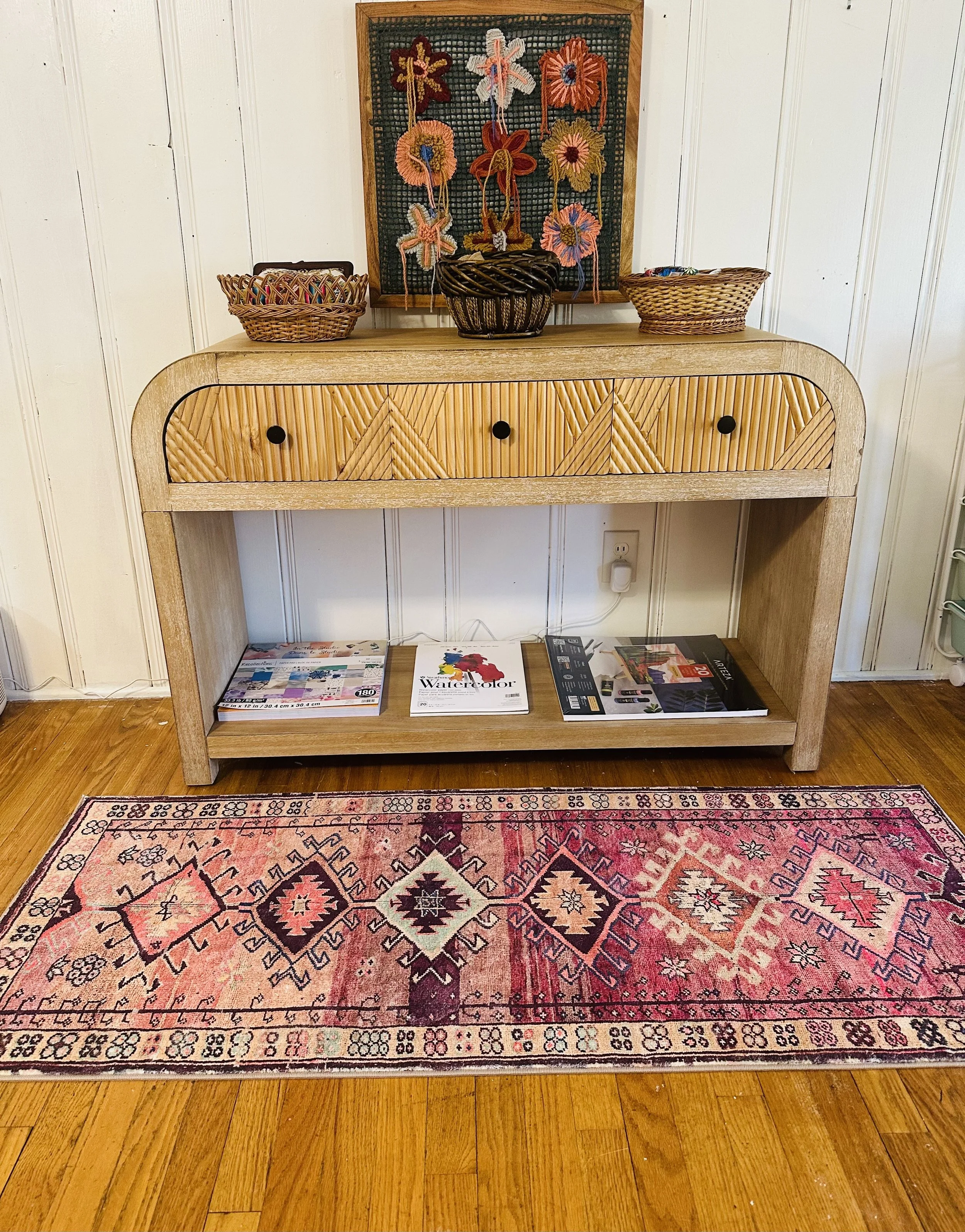 A wooden console table with a woven pattern, three drawers with black knobs, placed against a white paneled wall. On top of the table are three wicker baskets and a decorated framed artwork with colorful 3D floral designs. Below the table, on the woo
