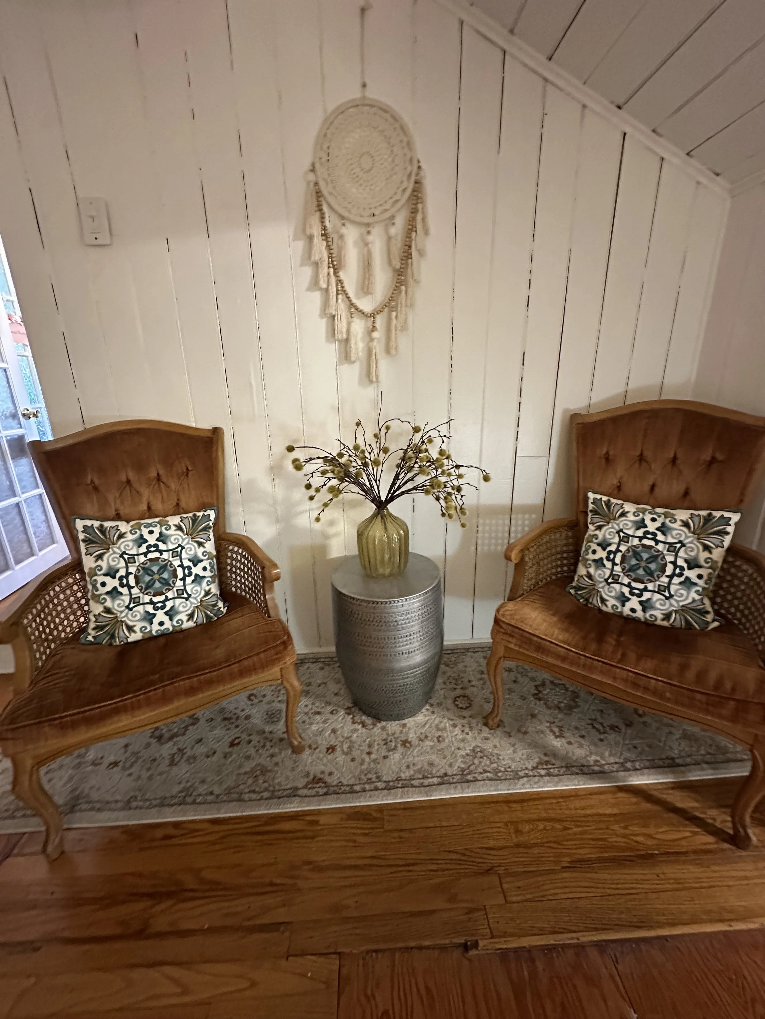 Two vintage armchairs with patterned pillows, a gray ceramic side table with a yellow vase and a gold-colored floral arrangement, white shiplap walls, a hanging dreamcatcher, and a window to the left.
