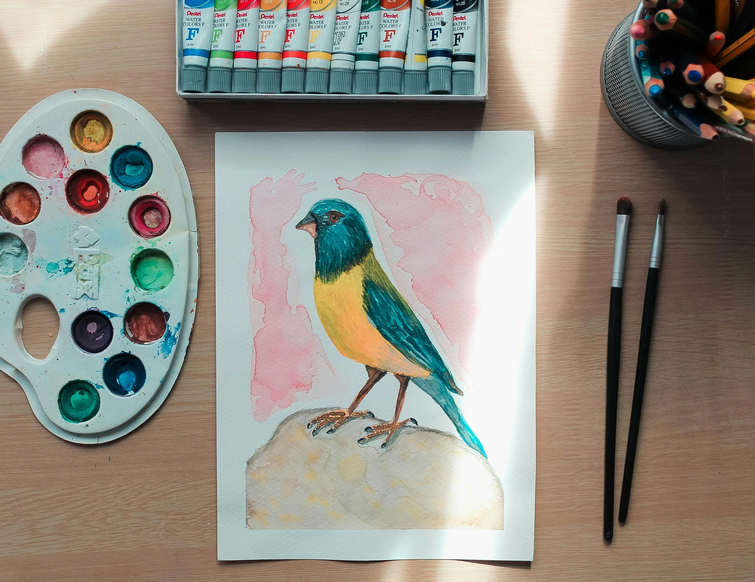 Watercolor painting of a colorful bird with yellow, blue, and green feathers, standing on a rock, on a wooden table with watercolor supplies nearby.
