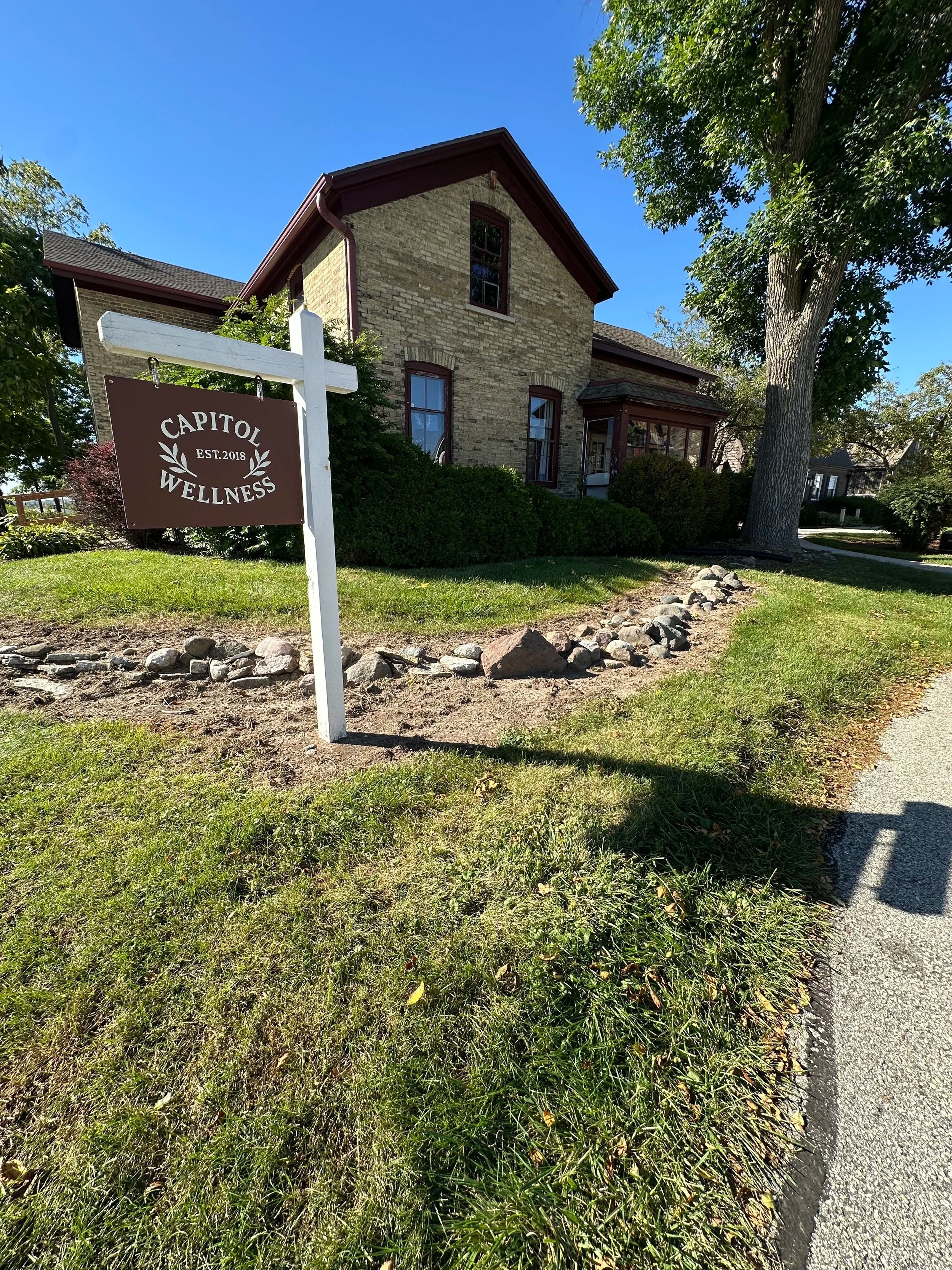 A house with brick walls, large windows, and a sloped roof, surrounded by trees and bushes, with a lawn and a rock-bordered flower bed in front. There is a sign that reads "Capitol Wellness" with "Est. 2018" on it, in the foreground.