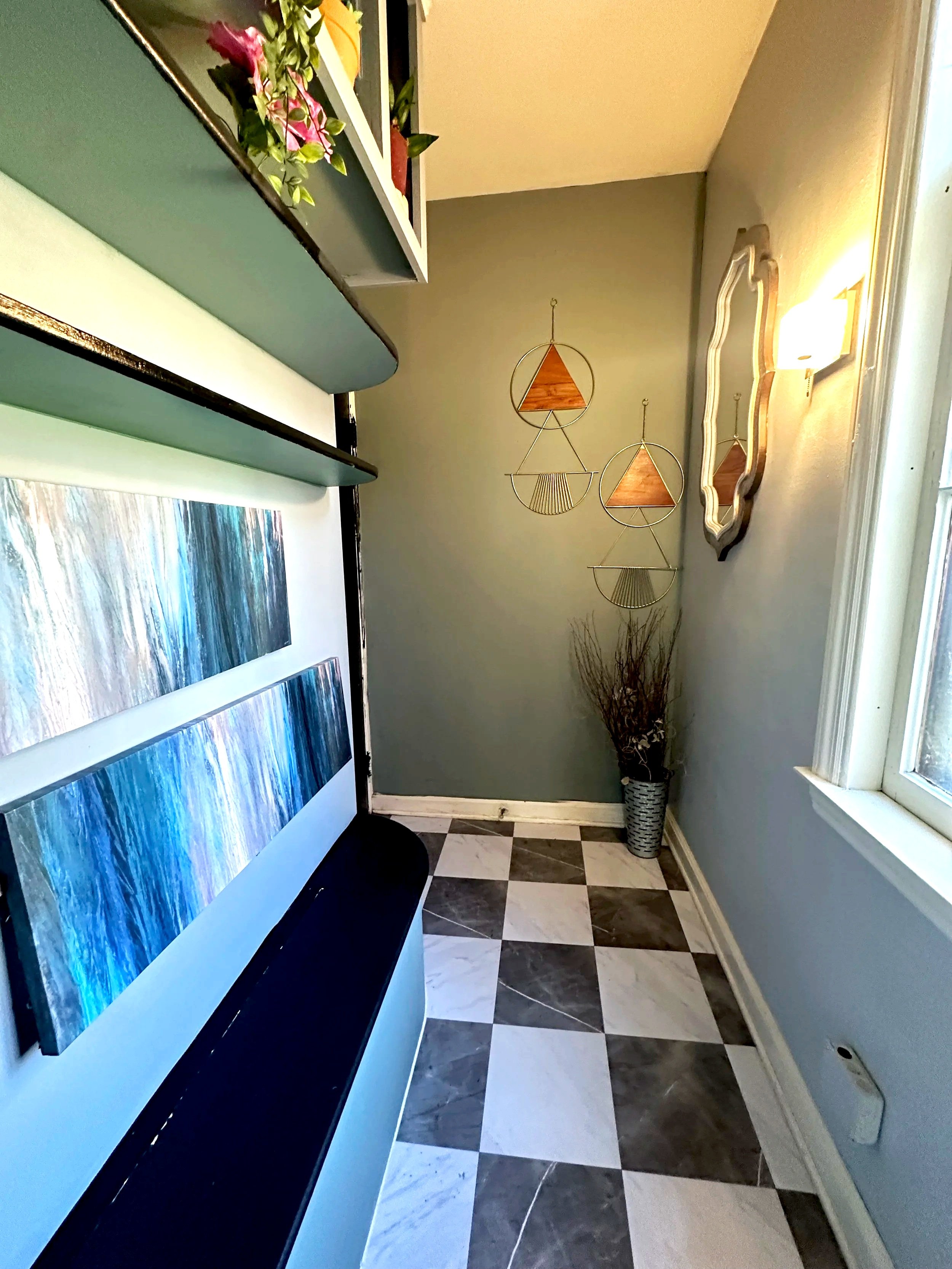 A corner of a room with a window on the right, a gray wall with wall art on the back wall, a mirror on the right wall, and black and white checkered flooring. There are shelves at the top left corner with decorations and an abstract painting below.