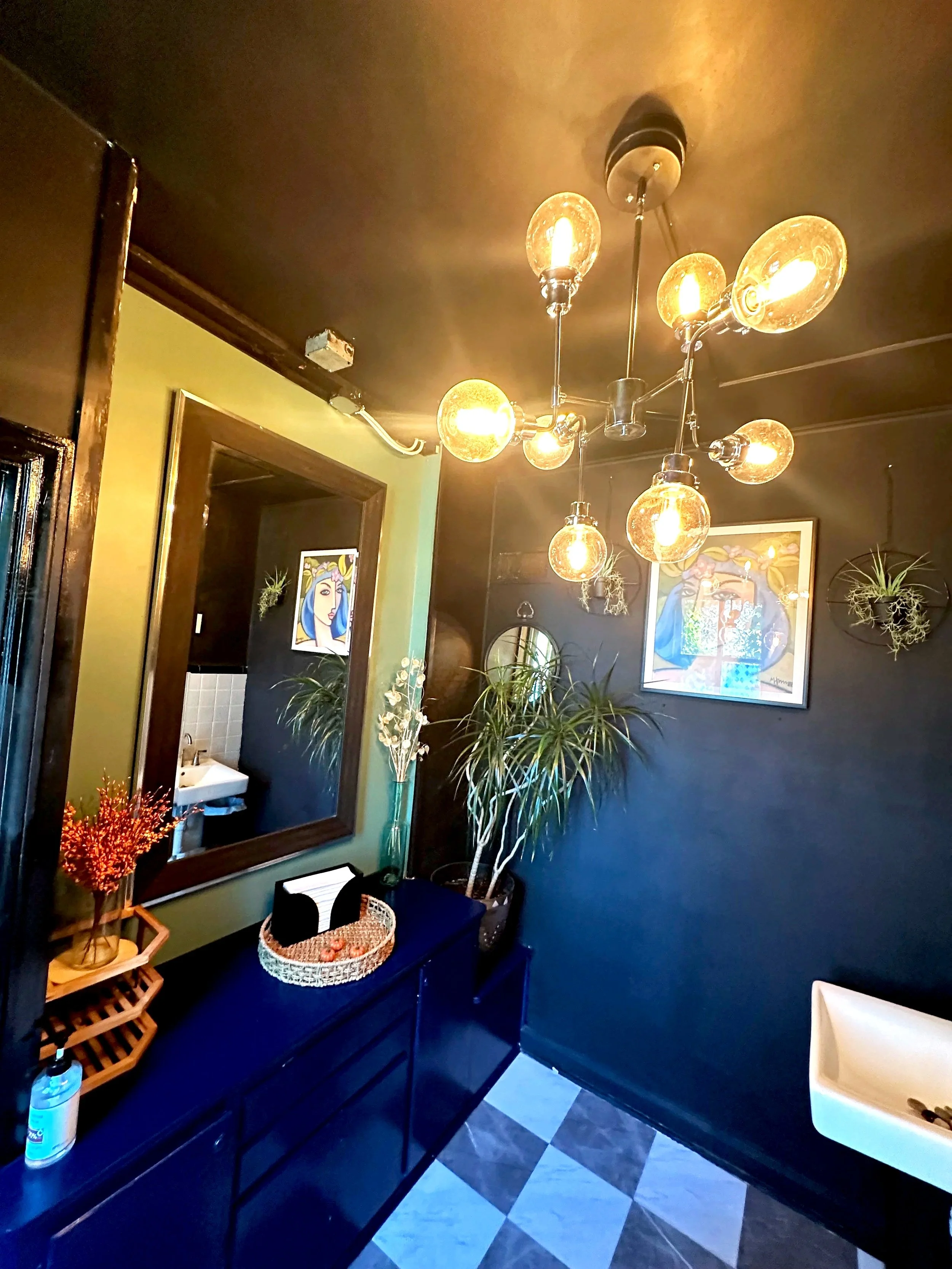 Interior of a room with a modern chandelier, potted plants, framed artwork, a mirror, a white wall-mounted sink, and a checkered floor.