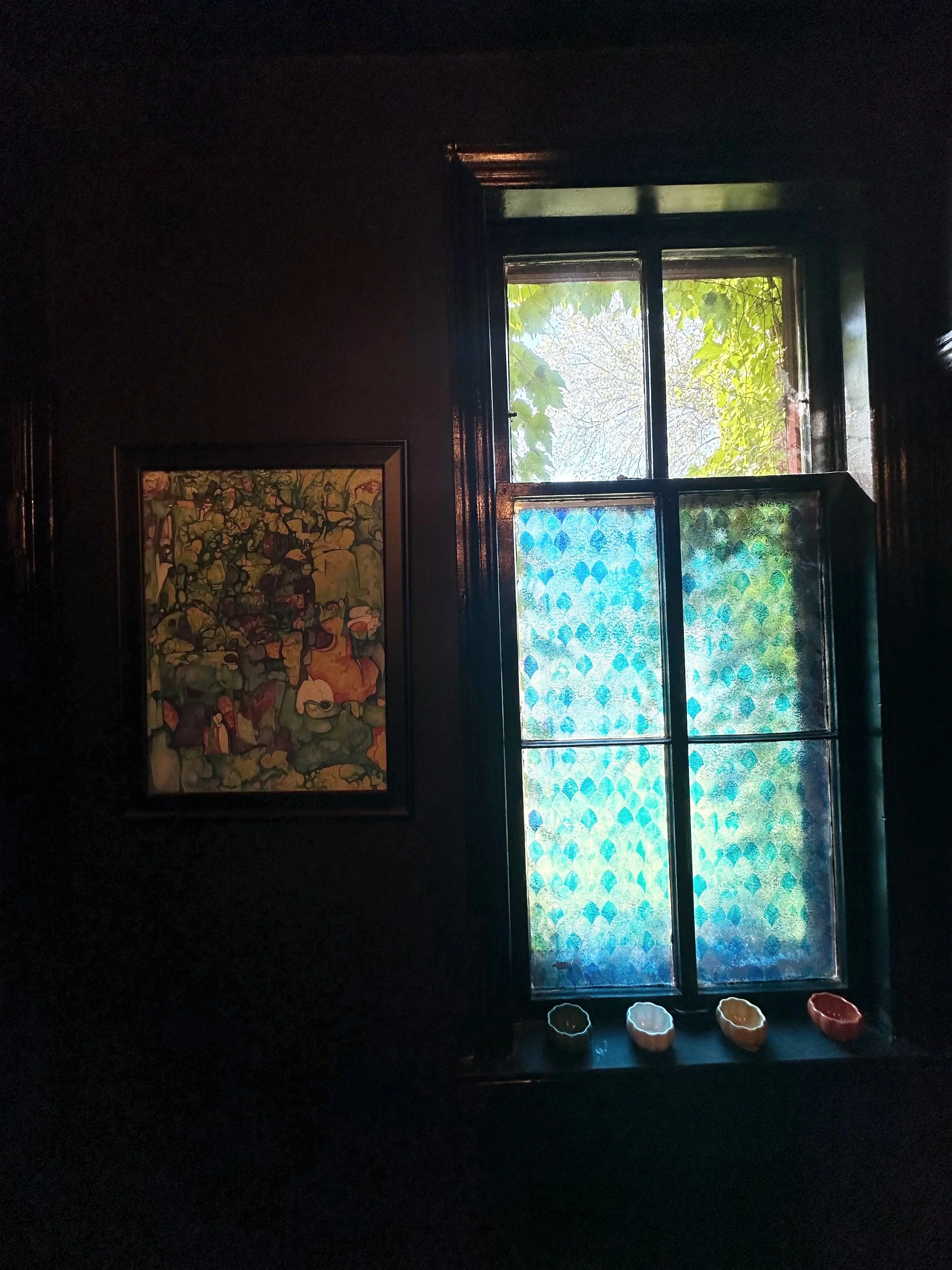 Dark interior room with a window showing green leaves outside. Four colorful ceramic bowls are on the windowsill. A framed abstract painting is hung on the wall beside the window.