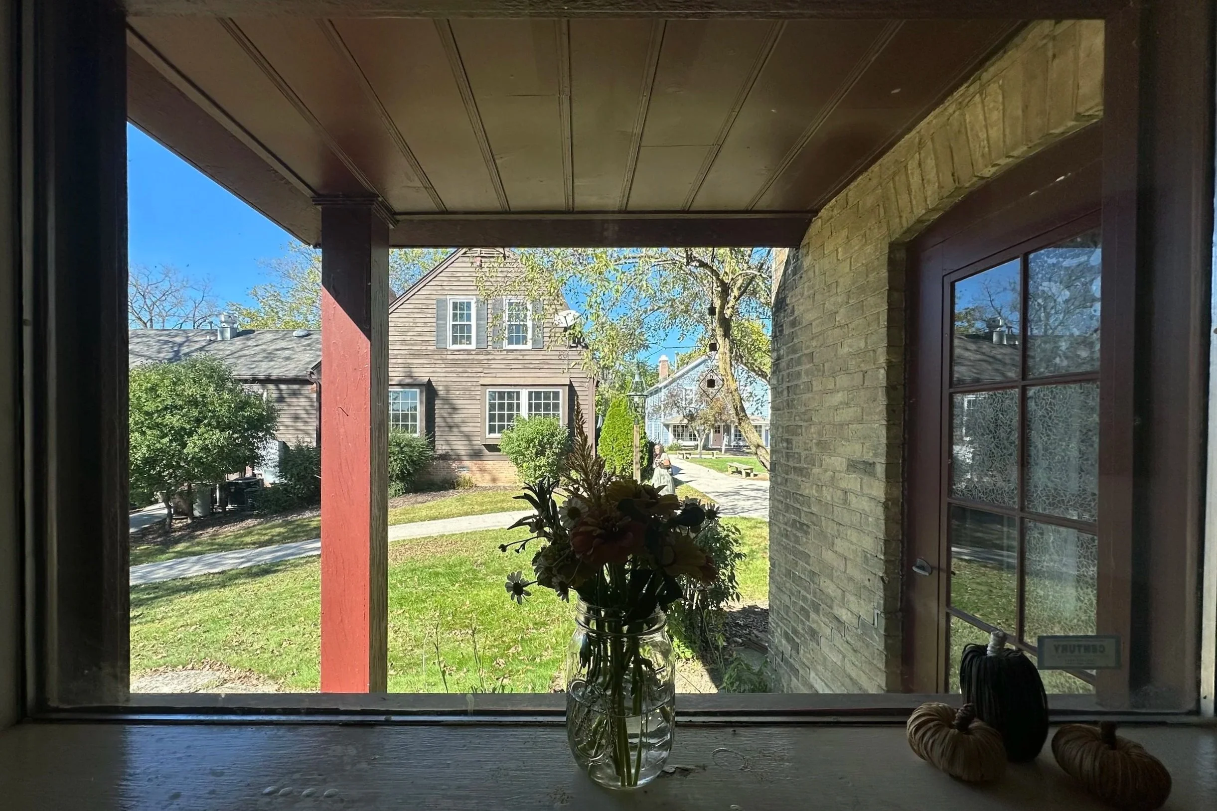 View through a window onto a residential street with houses, trees, and a sunny sky. Inside the window sill, there is a glass jar with a bouquet of flowers and two small pumpkins.
