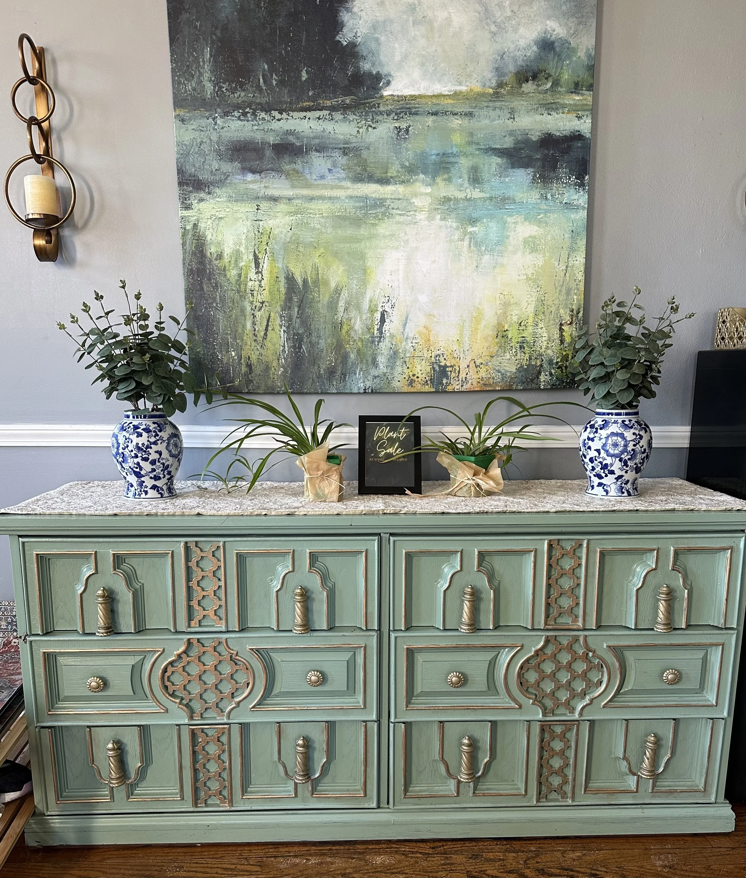 A vintage mint green wooden sideboard with ornate detailing and brass handles, topped with a white lace runner, topped with potted plants in blue and white porcelain vases and a small black framed sign that reads 'Plant Sale,' placed in front of an a