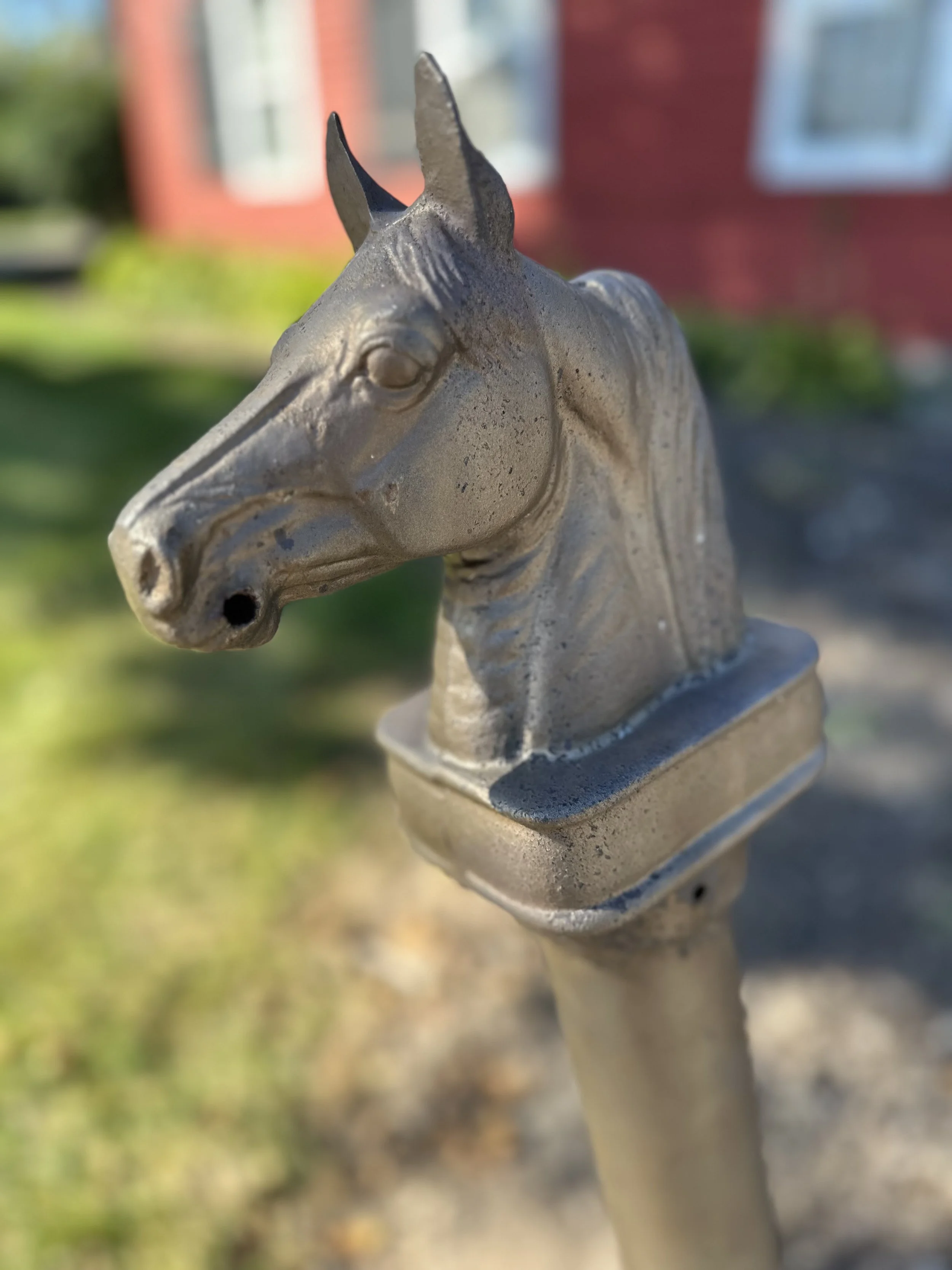 Close-up of a metallic horse head sculpture mounted on a pole outdoors, with a blurred background of a red building and greenery.