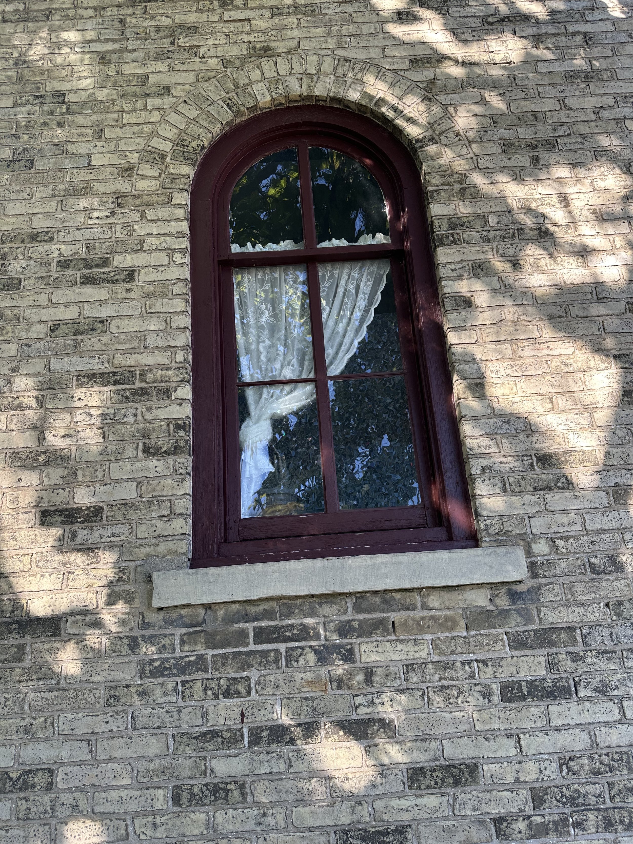 A brick building with a tall, arched window framed in dark red, with a white granite sill. The window reveals a white lace curtain and an outside view of green leaves. Shadows of trees are cast on the brick wall.