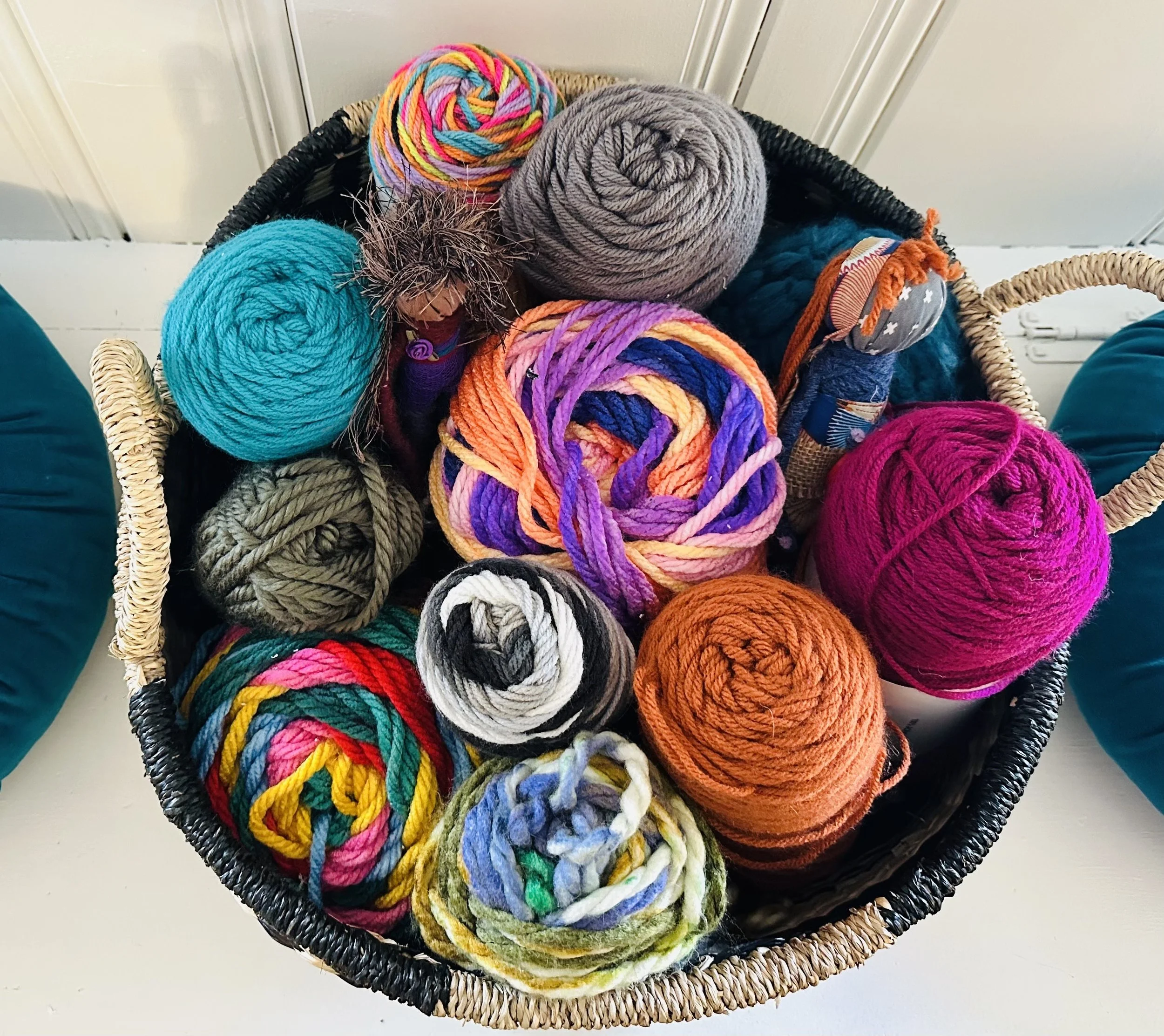 A black basket filled with multicolored skeins of yarn, some slim and others bulky, with textures that include smooth, fuzzy, and fluffy; the basket is on a white surface against a white wooden wall.