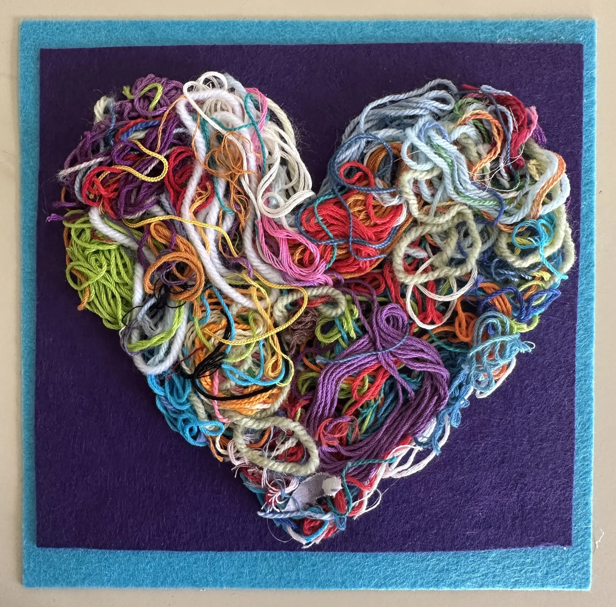 Colorful tangled embroidery floss arranged in the shape of a heart on a dark purple background with a light blue border.