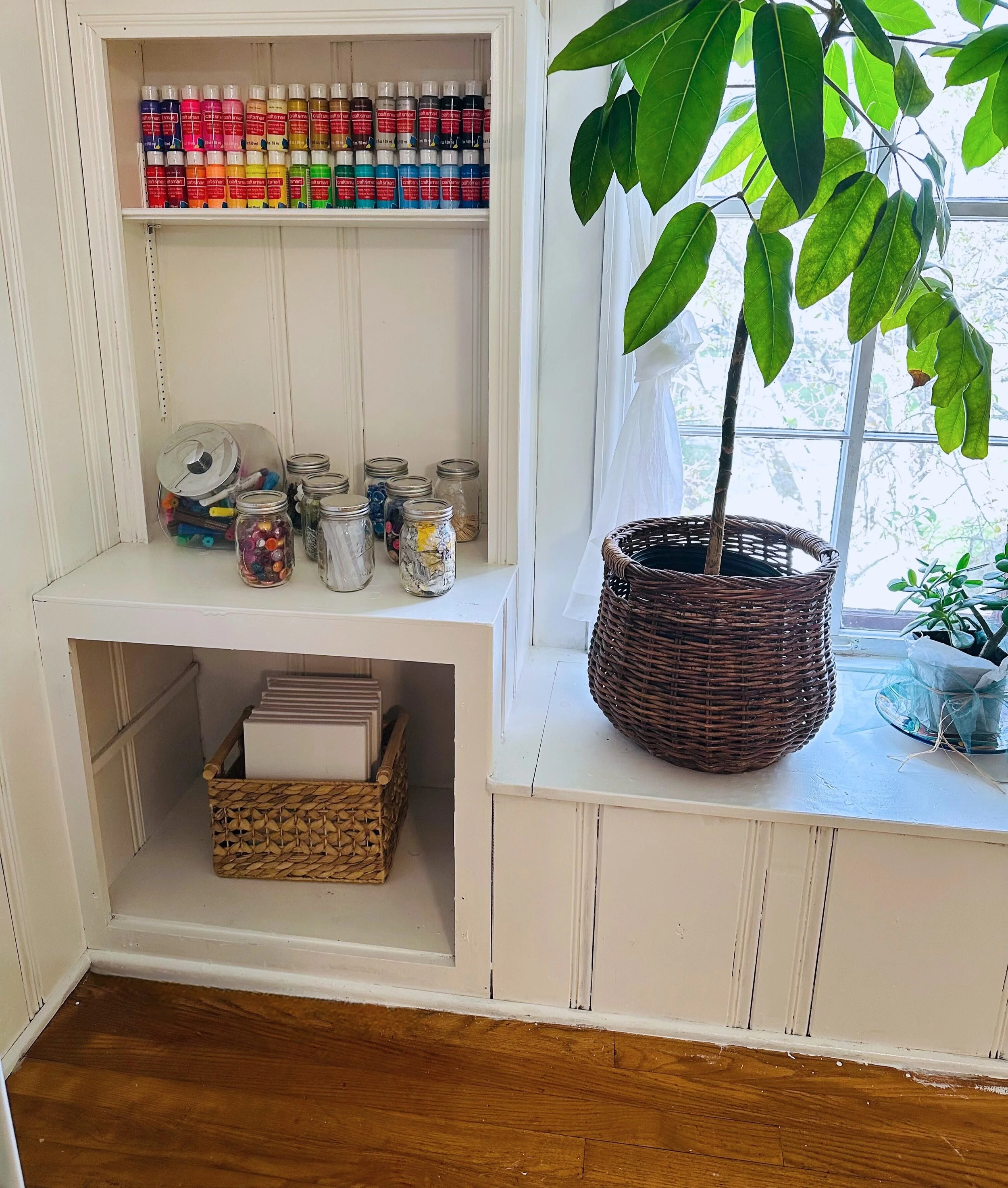 A built-in white wooden shelf unit with jars of crafts supplies, a small basket of white notebooks, a potted plant with large green leaves, and a wicker plant basket by a window with white curtains.