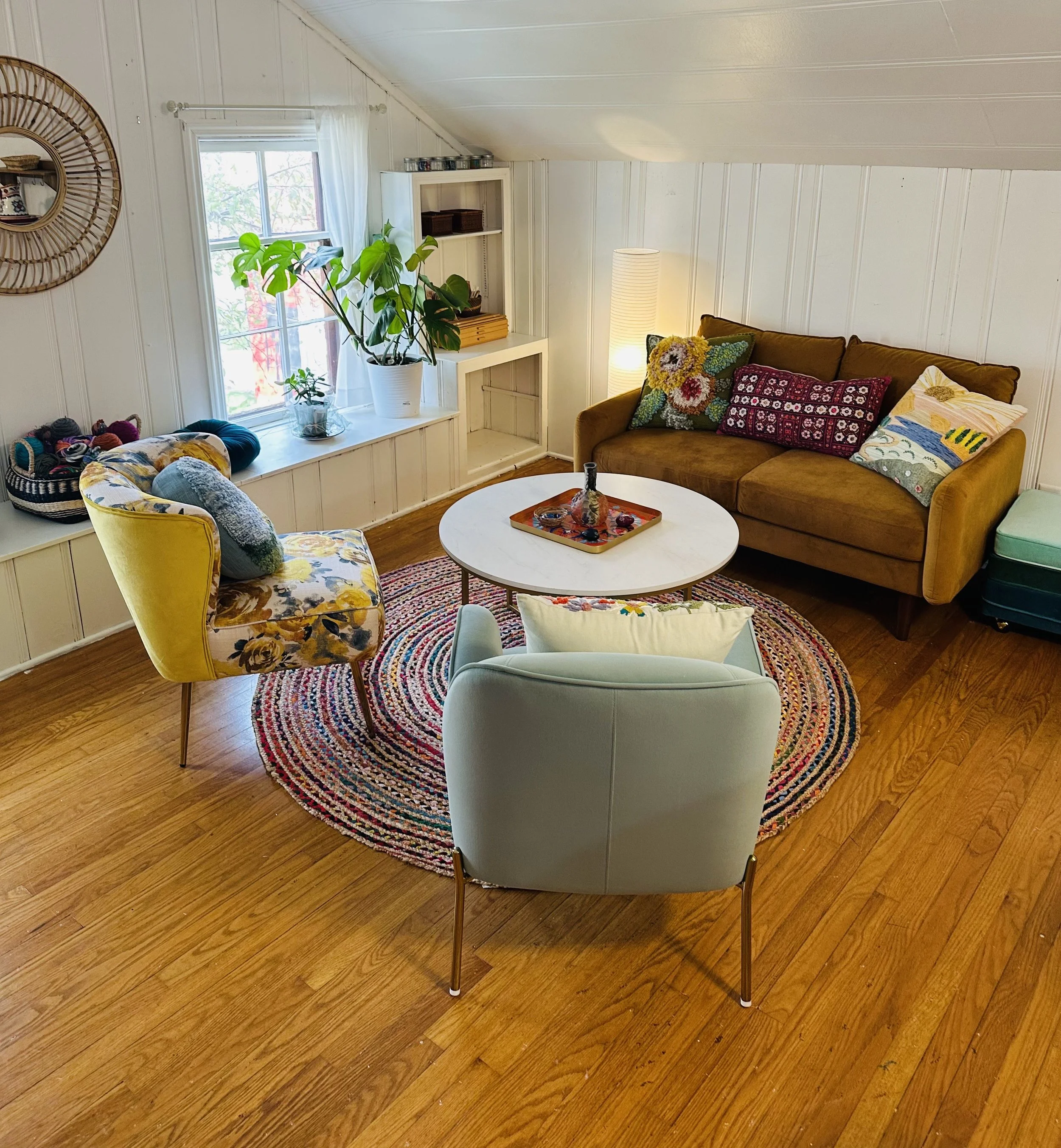 Cozy room with mid-century modern furniture, including a yellow floral armchair, light blue armchair with a white pillow, brown velvet sofa with embroidered pillows, round braided rug, white coffee table with a tray, potted plants near a window, and 