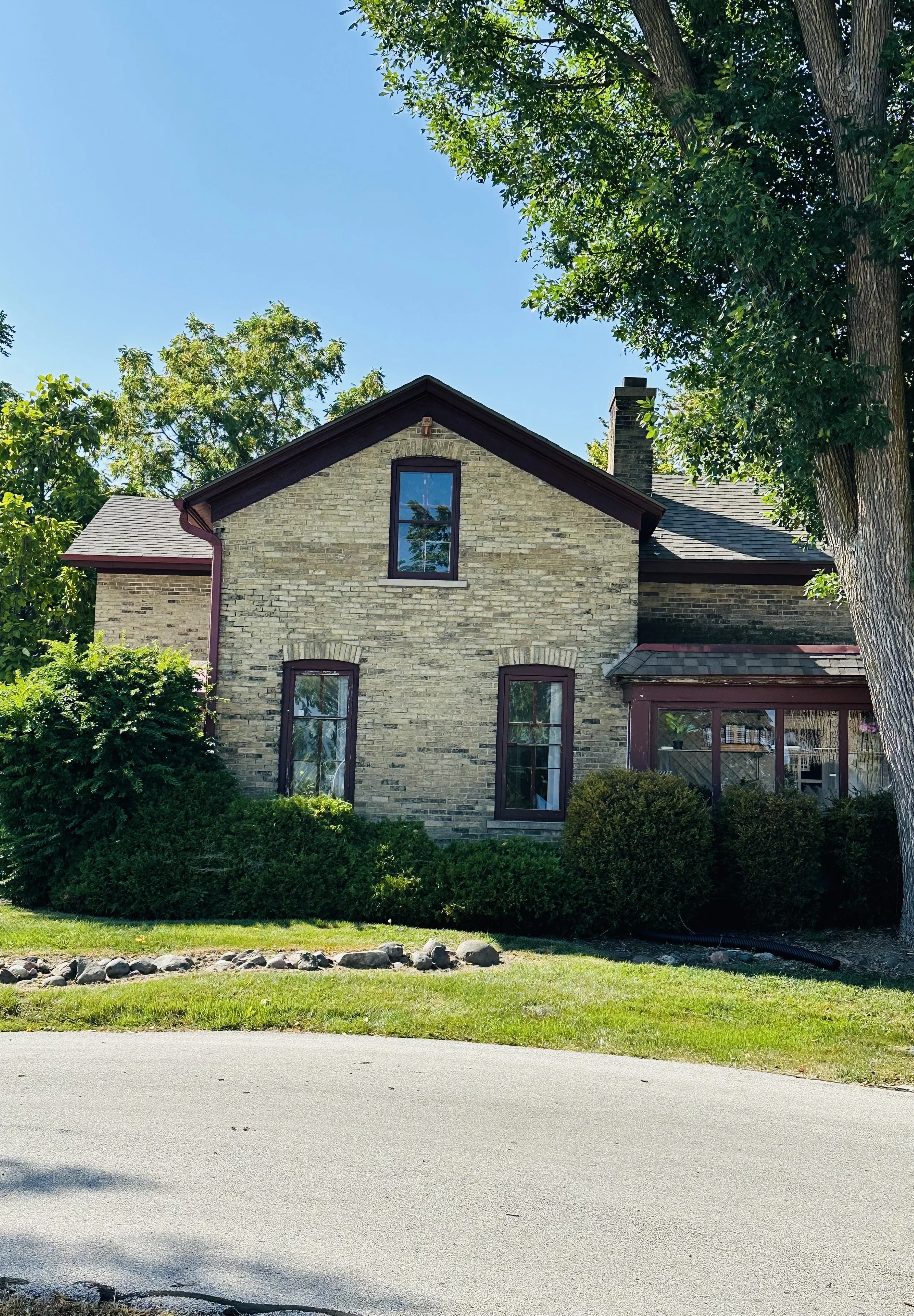 A two-story brick house with a gabled roof, surrounded by green bushes and trees under a clear blue sky.
