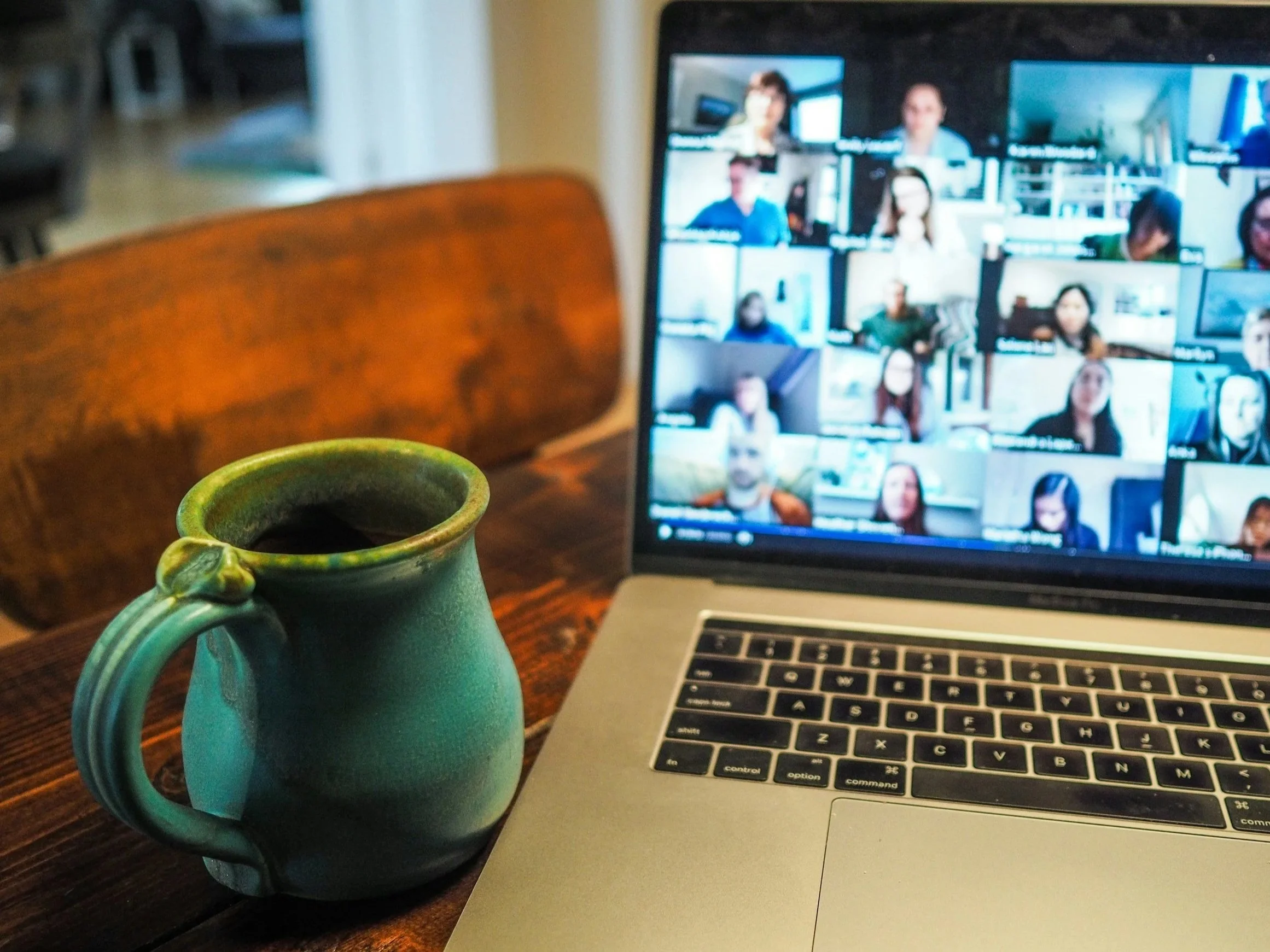 A ceramic mug filled with coffee, placed on a wooden table next to an open laptop showing a video conference with multiple participants in a grid layout.
