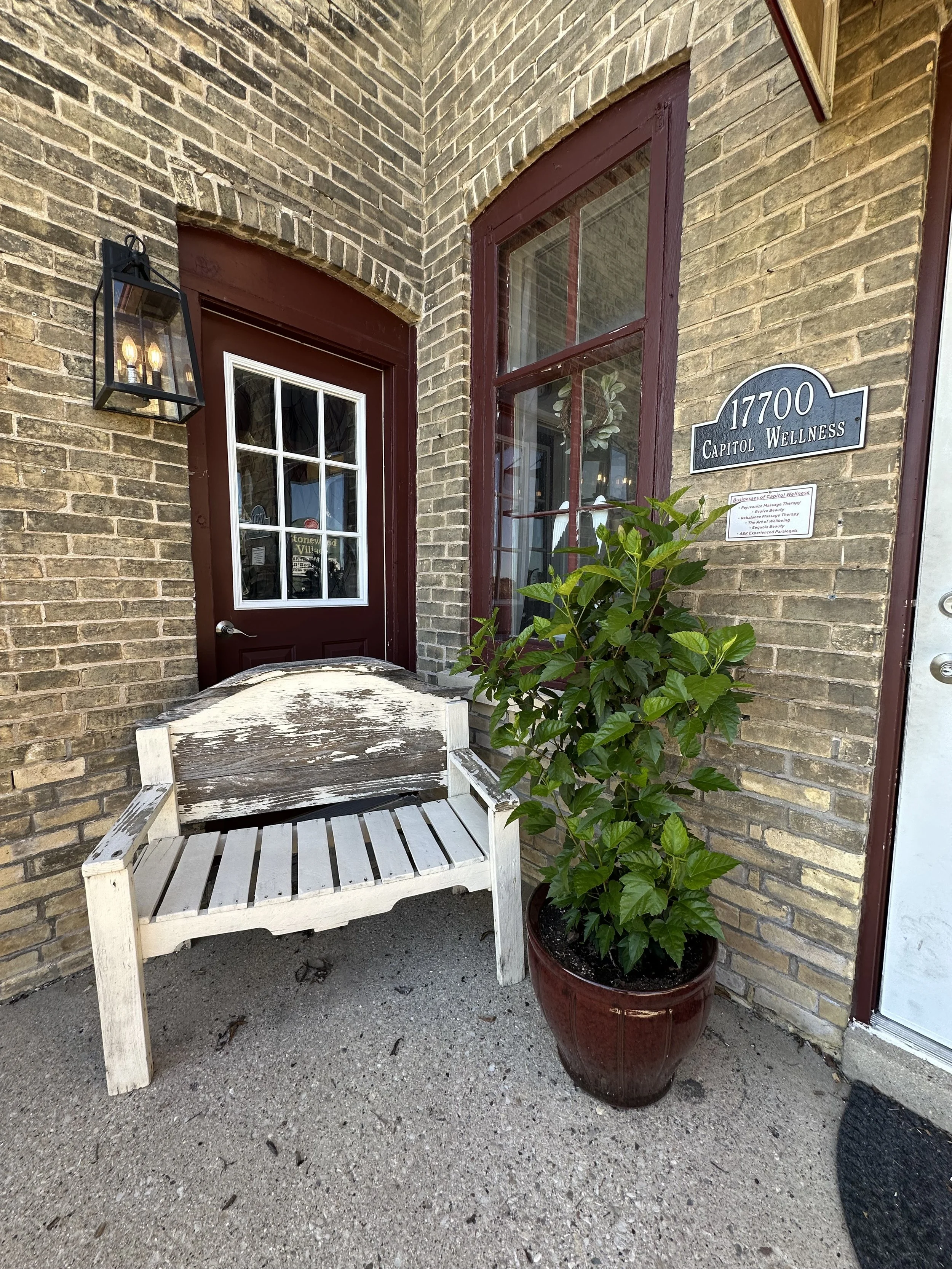Outdoor scene showing a brick building with a burgundy door with a window, a white wooden bench with peeling paint, and a large potted green plant next to the door. There is a street address sign reading '17700 Capitol Wellness' and a black outdoor l
