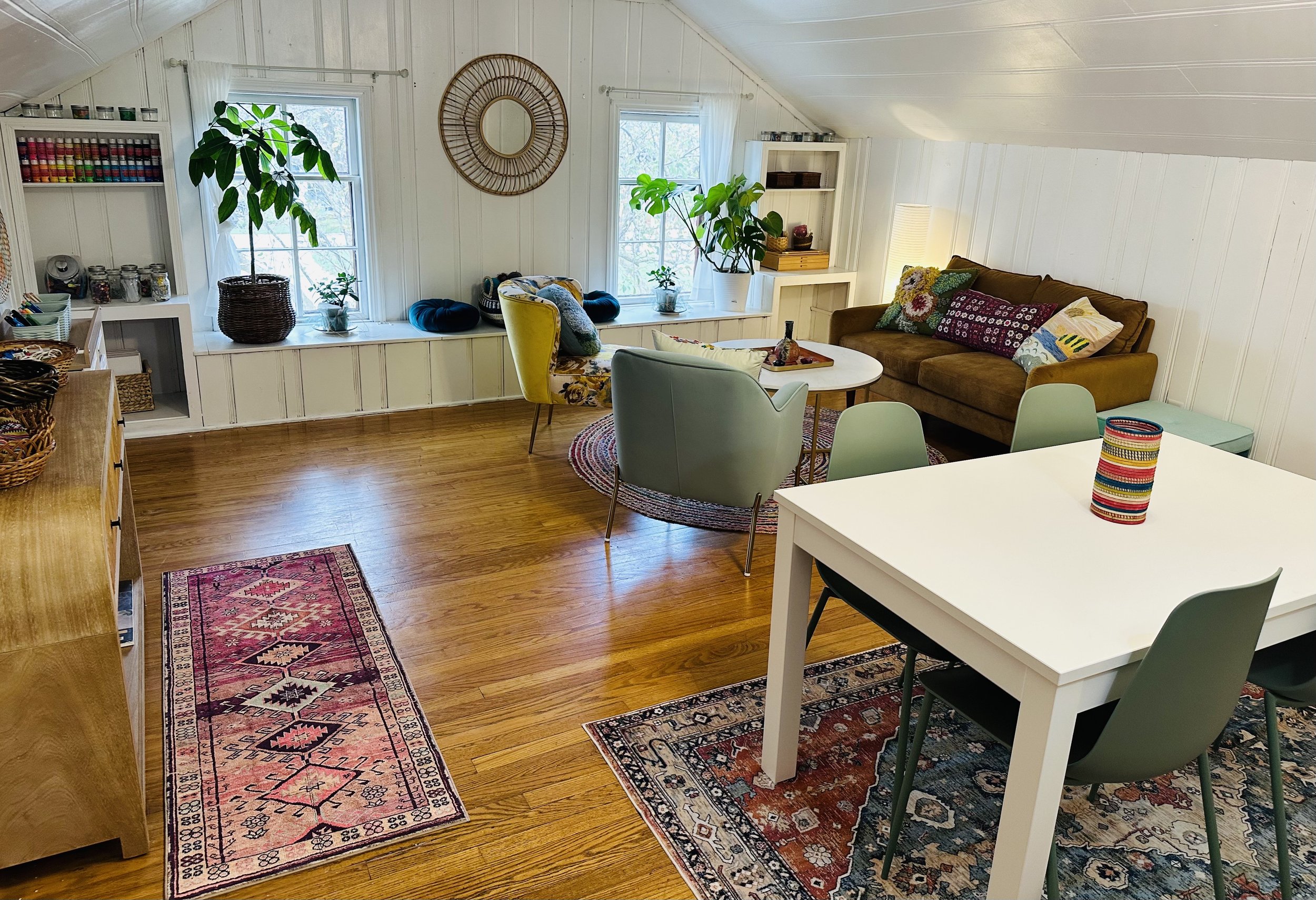 Cozy room with white wood-paneled walls, hardwood floors, and large windows with white curtains. Features a brown sofa with colorful pillows, a white round coffee table, a green armchair, and a patterned area rug. A white dining table with four chair