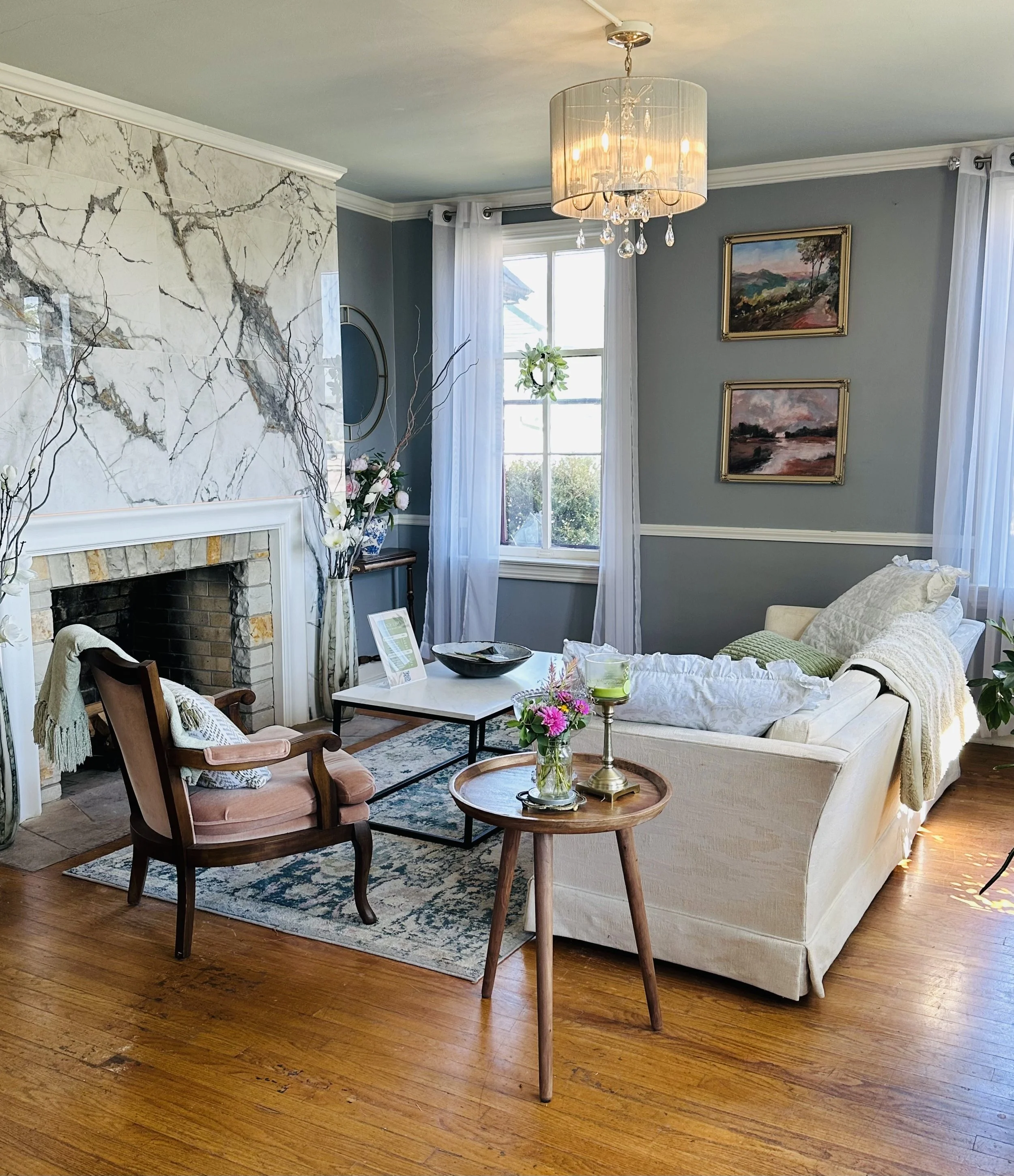 Living room with a fireplace, a white sofa, a wooden armchair, a coffee table, and decor including paintings, flowers, and a chandelier.