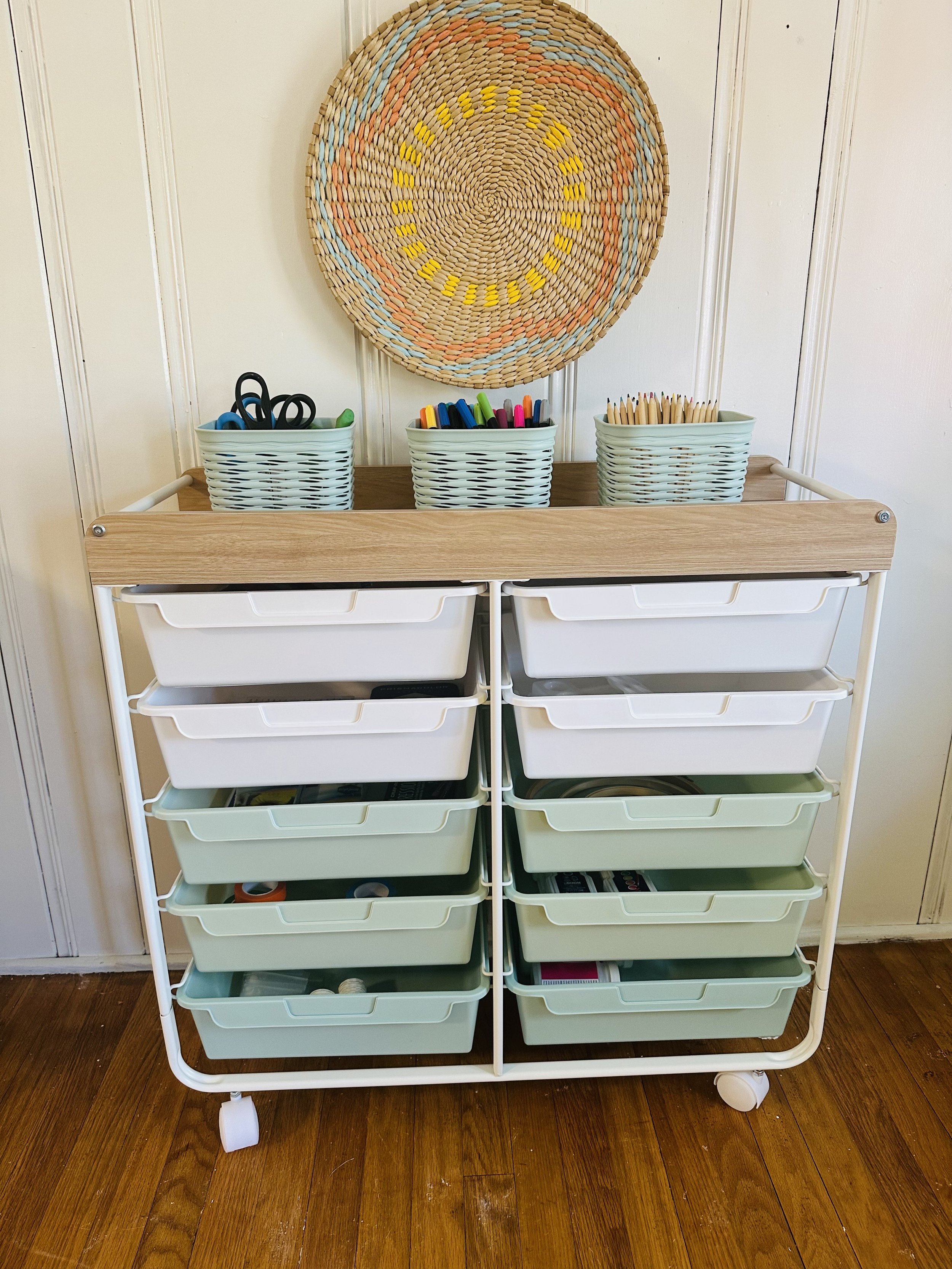 A craft storage cart with white and green plastic drawers, a wooden top, and basket containers holding scissors, markers, and colored pencils. A woven wall hanging decorates the wall behind it.