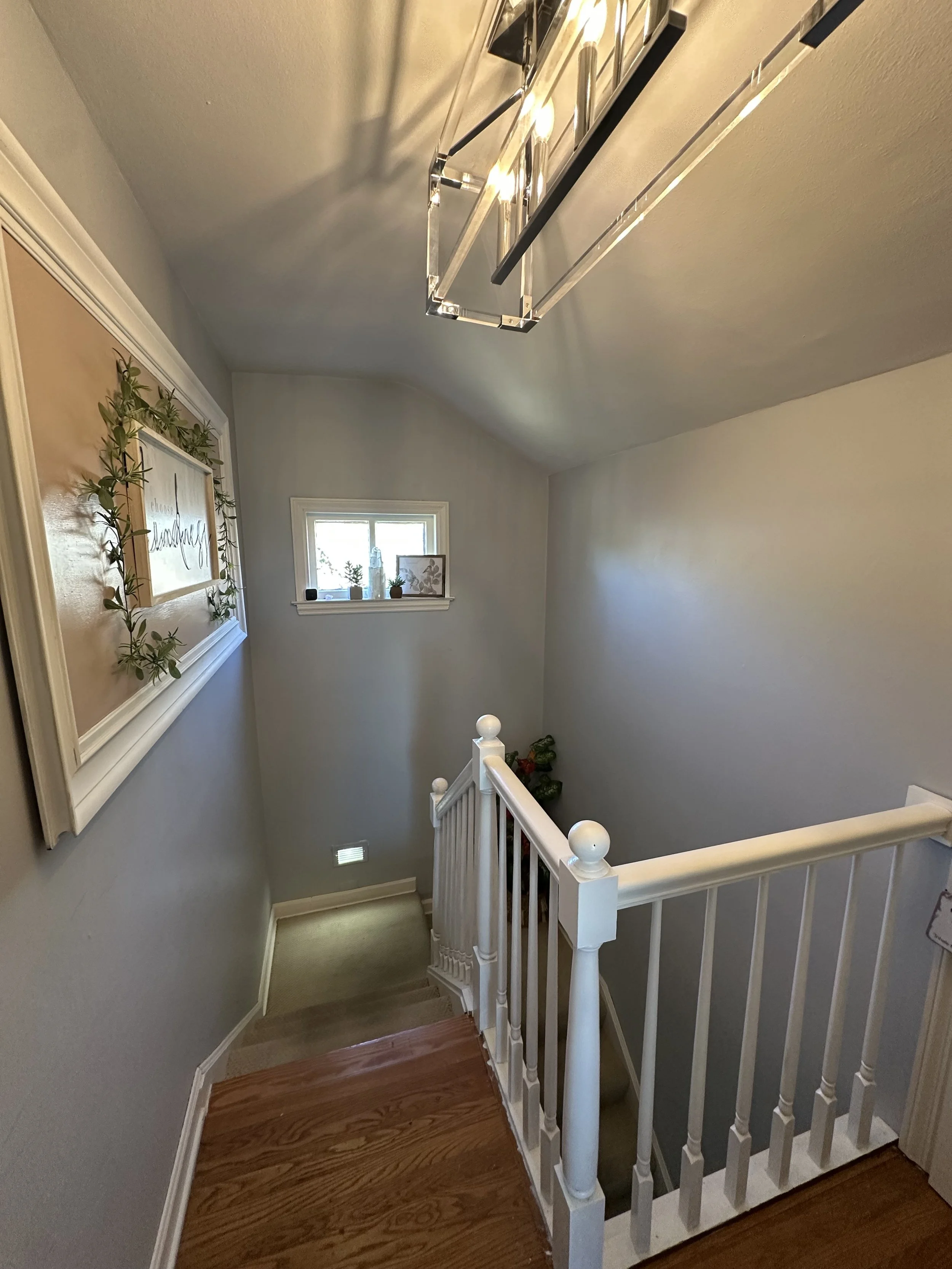 View from the top of a staircase looking down, with a white railing on the right and a small window at the end of the staircase. The ceiling has a modern rectangular light fixture, and the walls are decorated with a framed sign and plants.