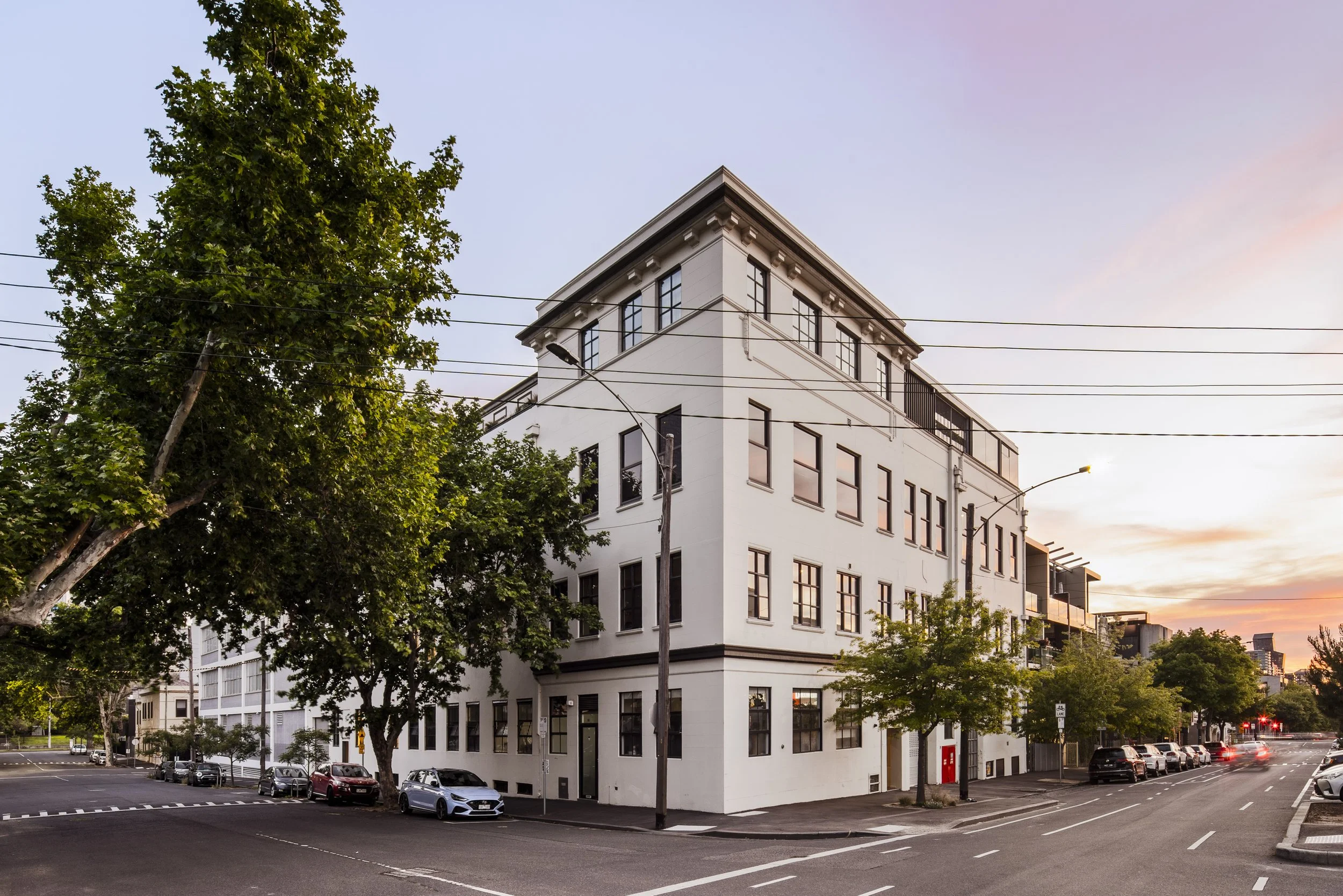 A white multi-story building on a city street corner with trees and parked cars, during sunset.