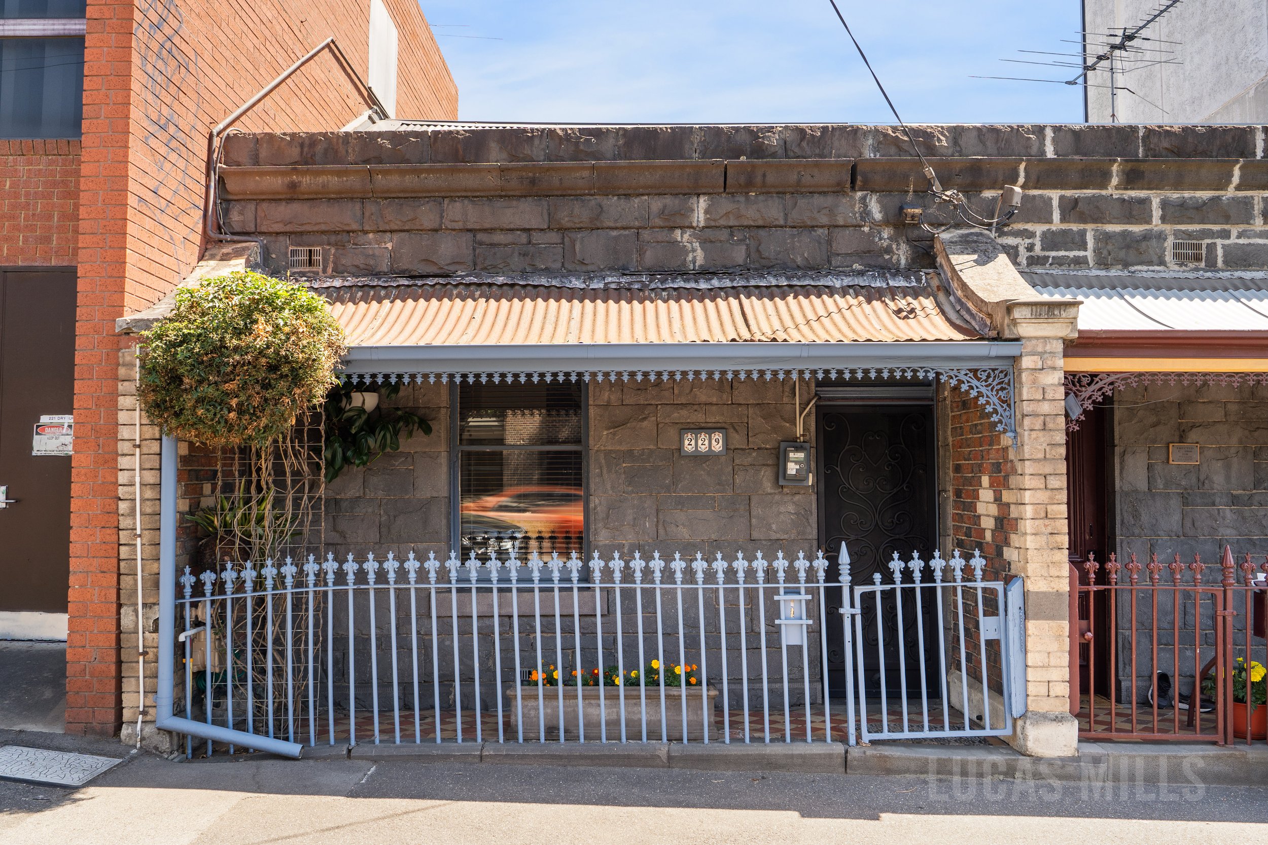 Urban courtyard with potted plants, a leafless tree, a glass door with a blue frame, a wicker outdoor sofa with colorful cushions, and potted plants along a brick and concrete wall.