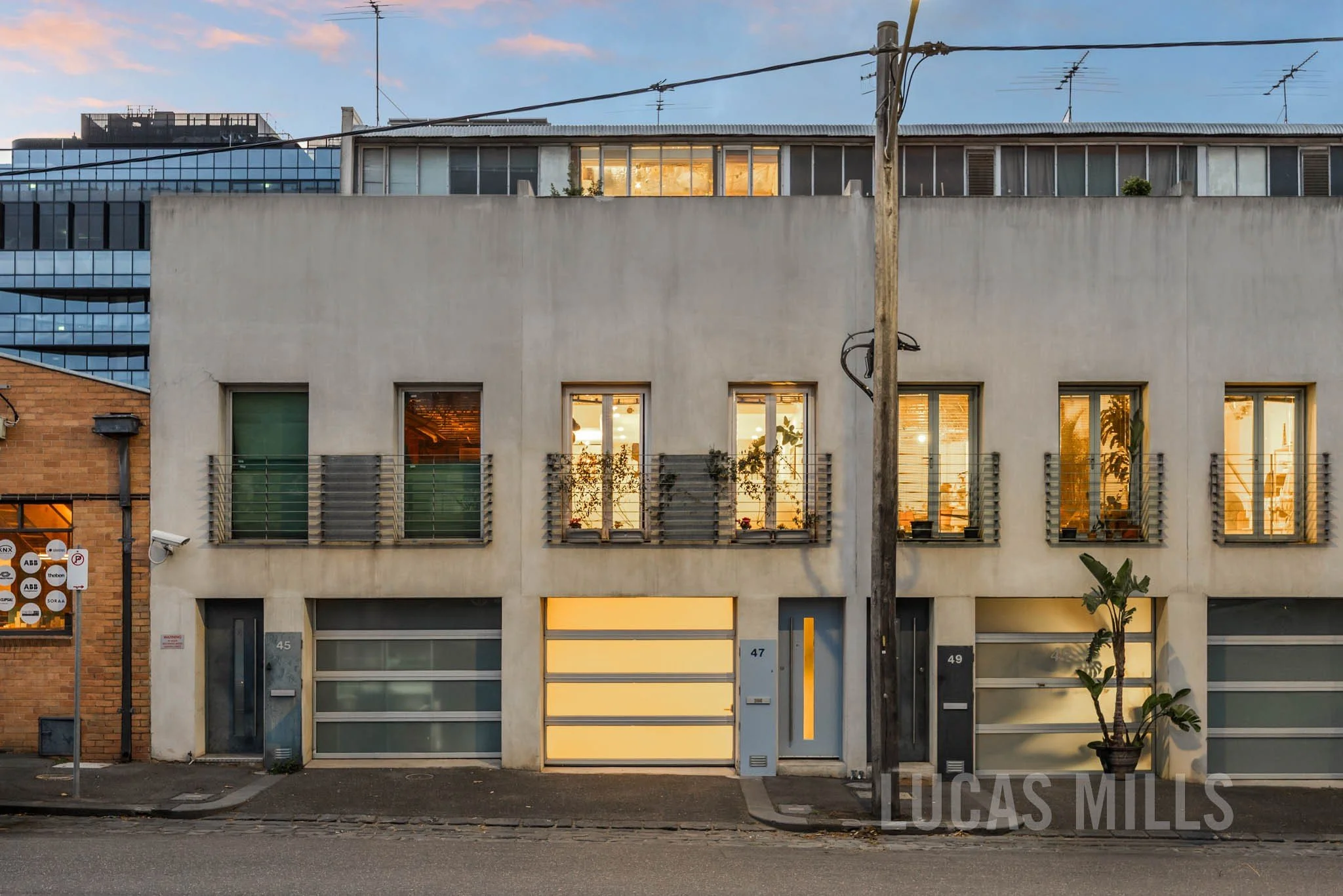 Modern urban building with three stories, featuring large windows with warm interior lighting, a garage door, and a small potted plant outside. An old utility pole is in front.