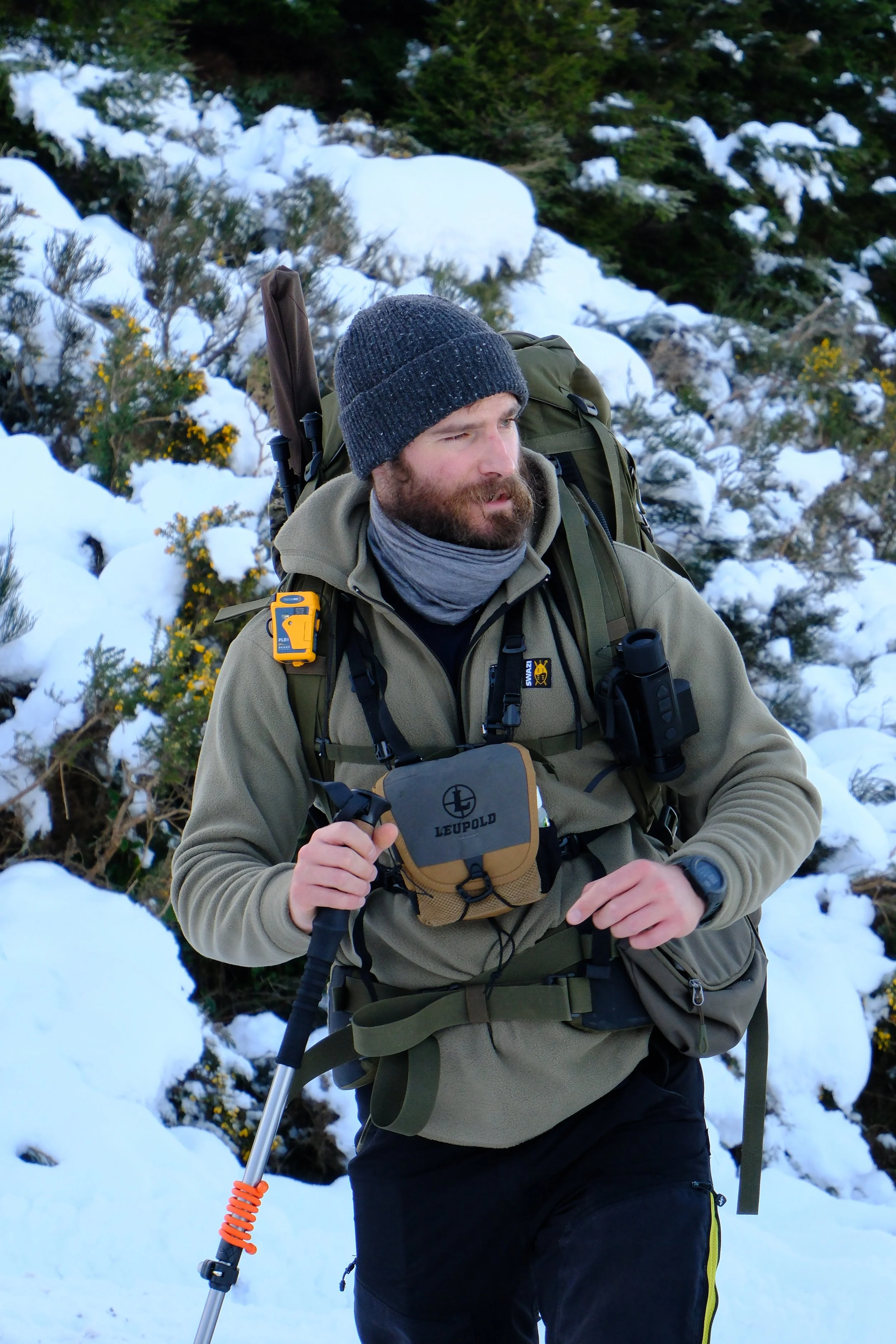 A man hiking outdoors in a snowy forest, wearing winter clothing, a beanie, carrying a backpack, and holding a trekking pole.