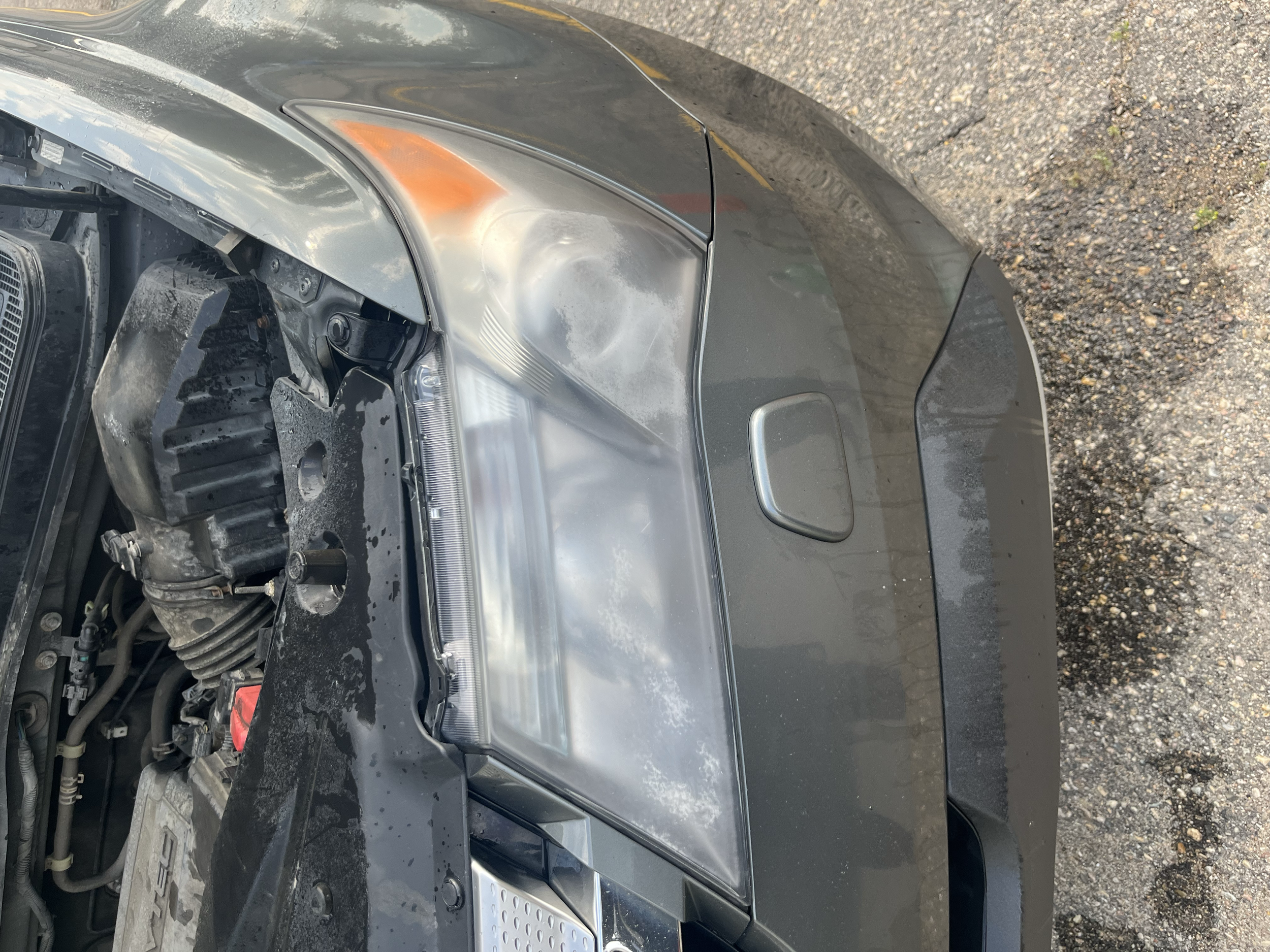 Close-up of a damaged car front with the hood open, headlight missing, visible engine components, and scratches on the surface, on a gravel road.