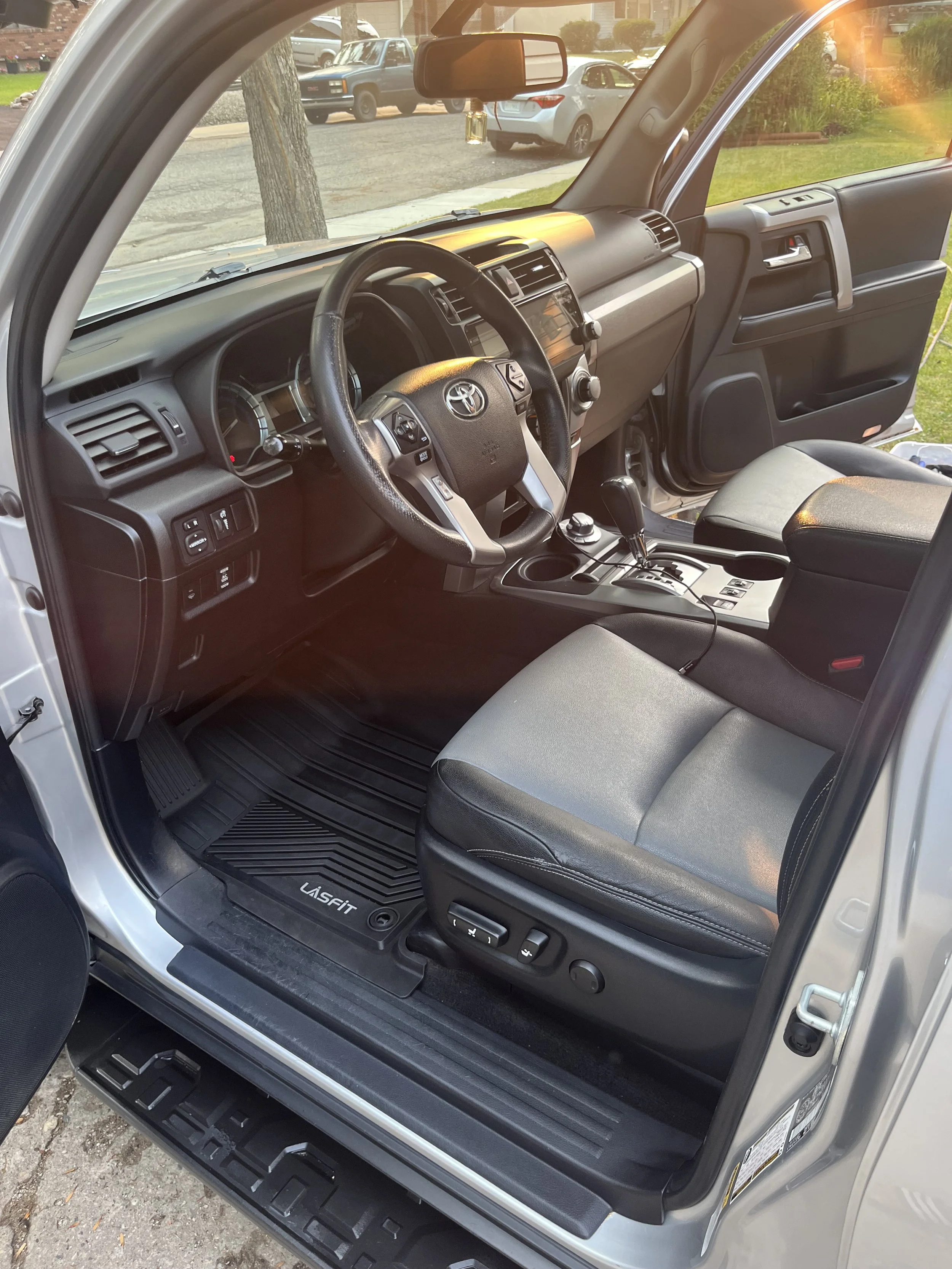 Interior of a Toyota vehicle showing the driver’s seat, steering wheel, dashboard, and center console with gear shift. The seat has controls visible, and the floor mat says 'LusFit.' Outside the vehicle, other parked cars and a tree are visible.