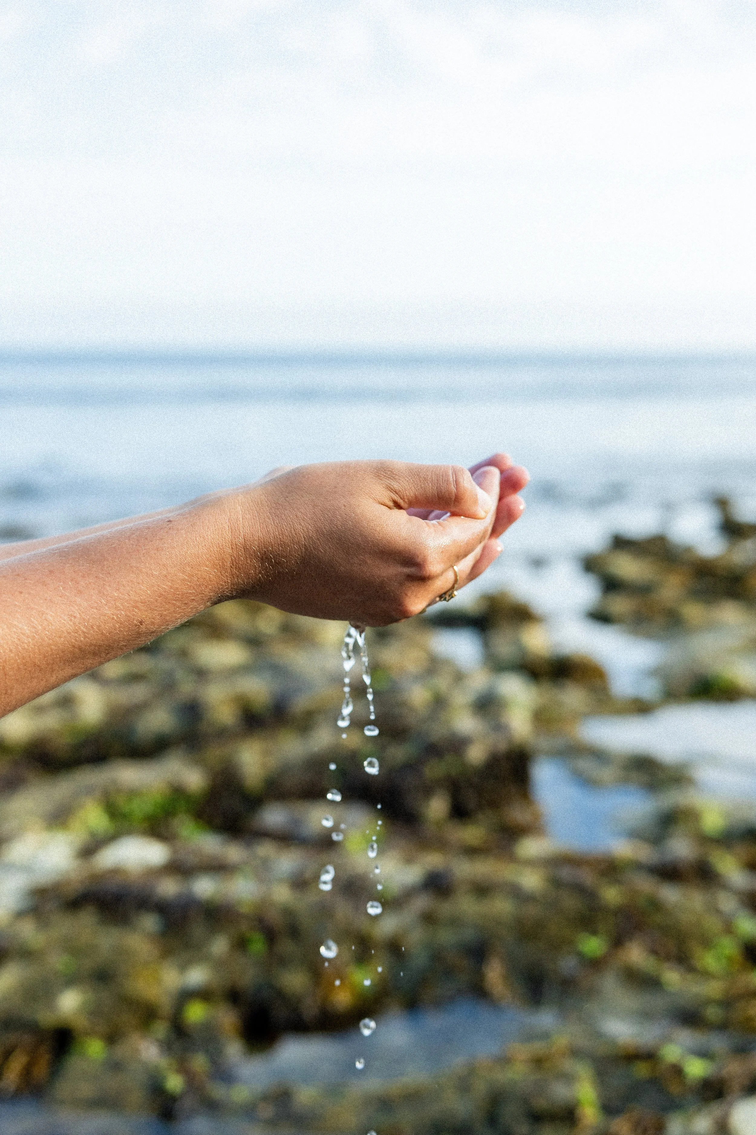 Close-up of a person’s hand releasing water droplets at the ocean shoreline with rocky background and horizon line in the distance.