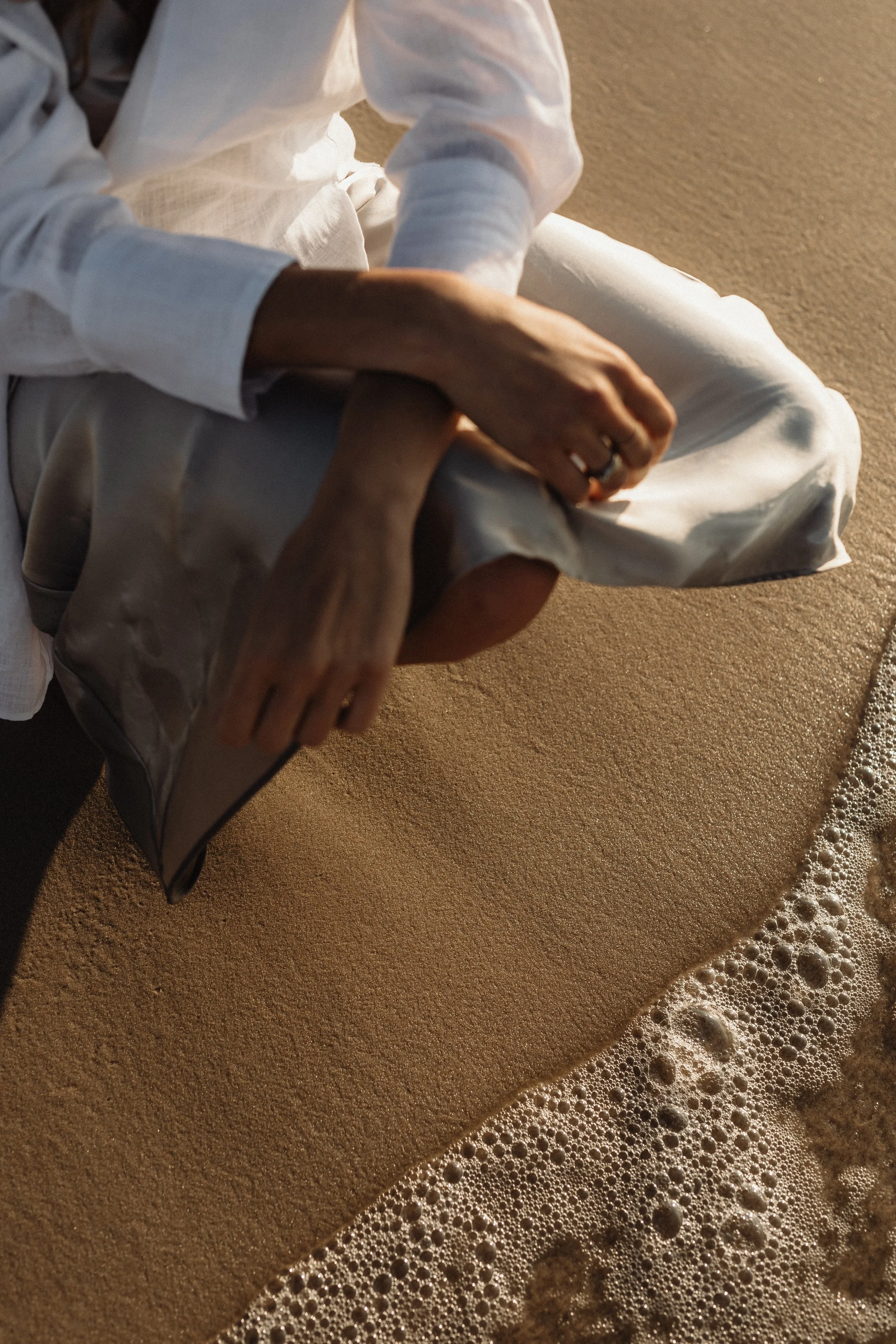 Close-up of a person wearing a white long-sleeve shirt and white pants, sitting on a sandy beach near the water's edge with foam and bubbles from the ocean visible.
