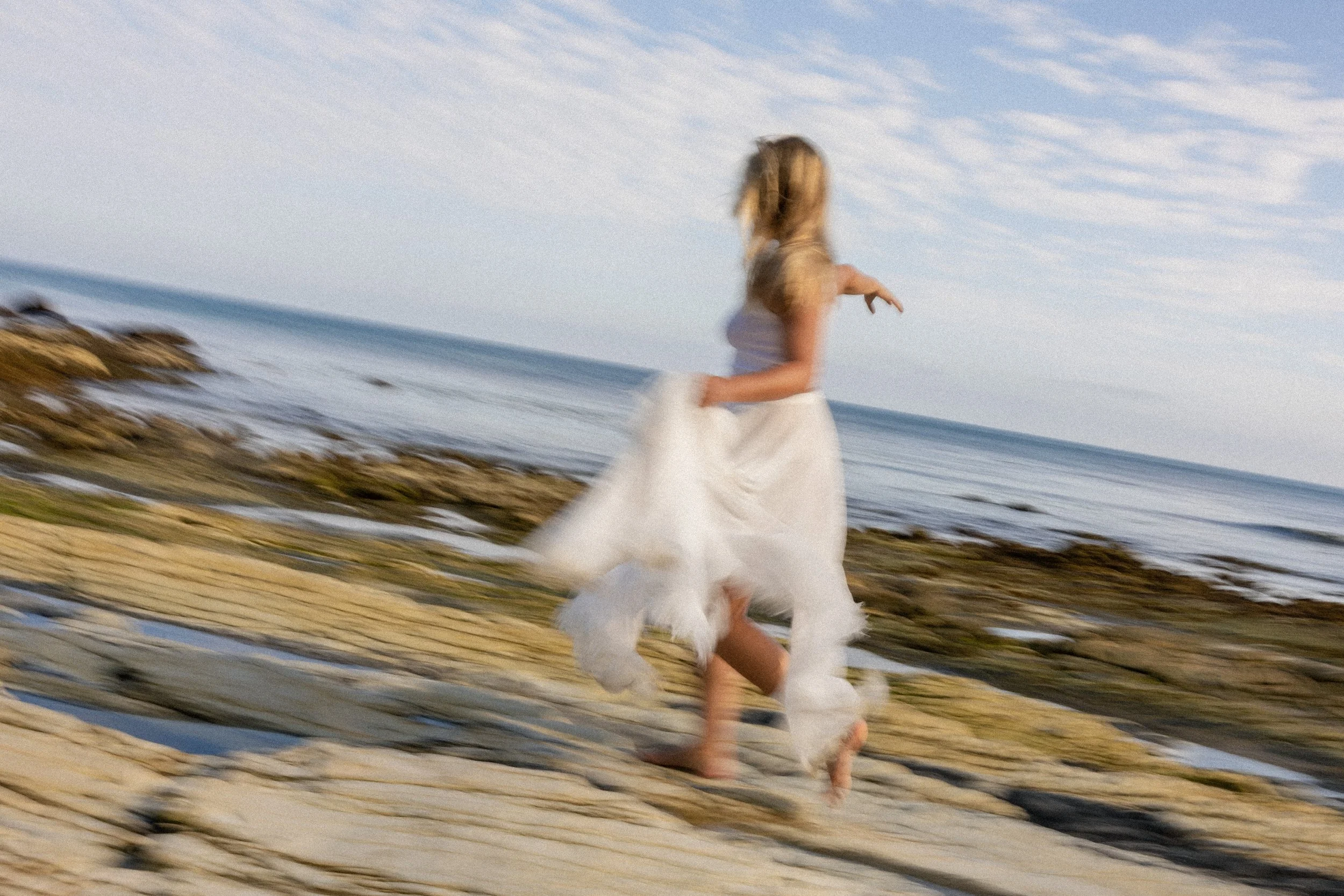 A woman in a white dress running along a rocky shoreline at the beach.