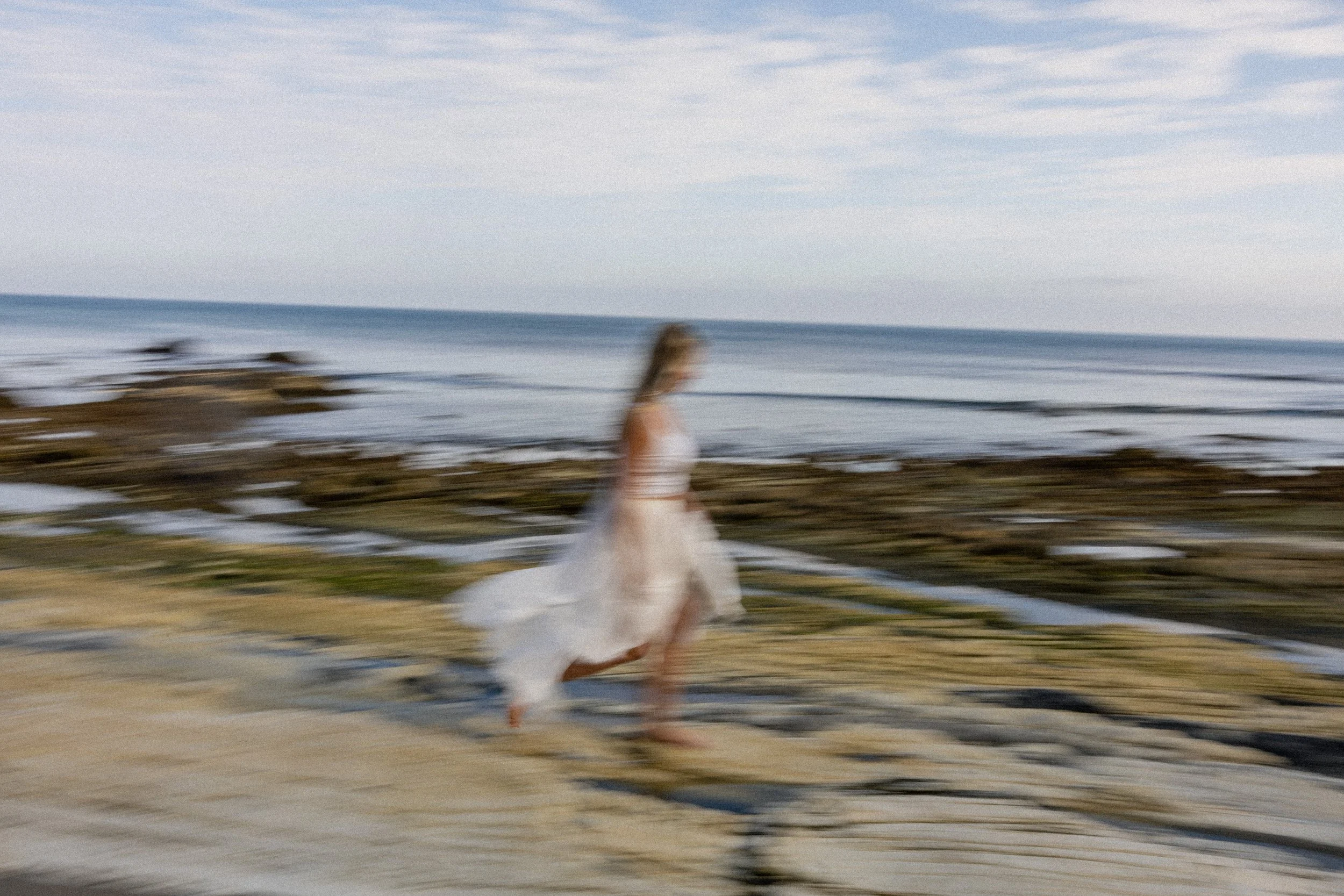 A blurred woman in a white dress running on a beach with water and sky in the background.