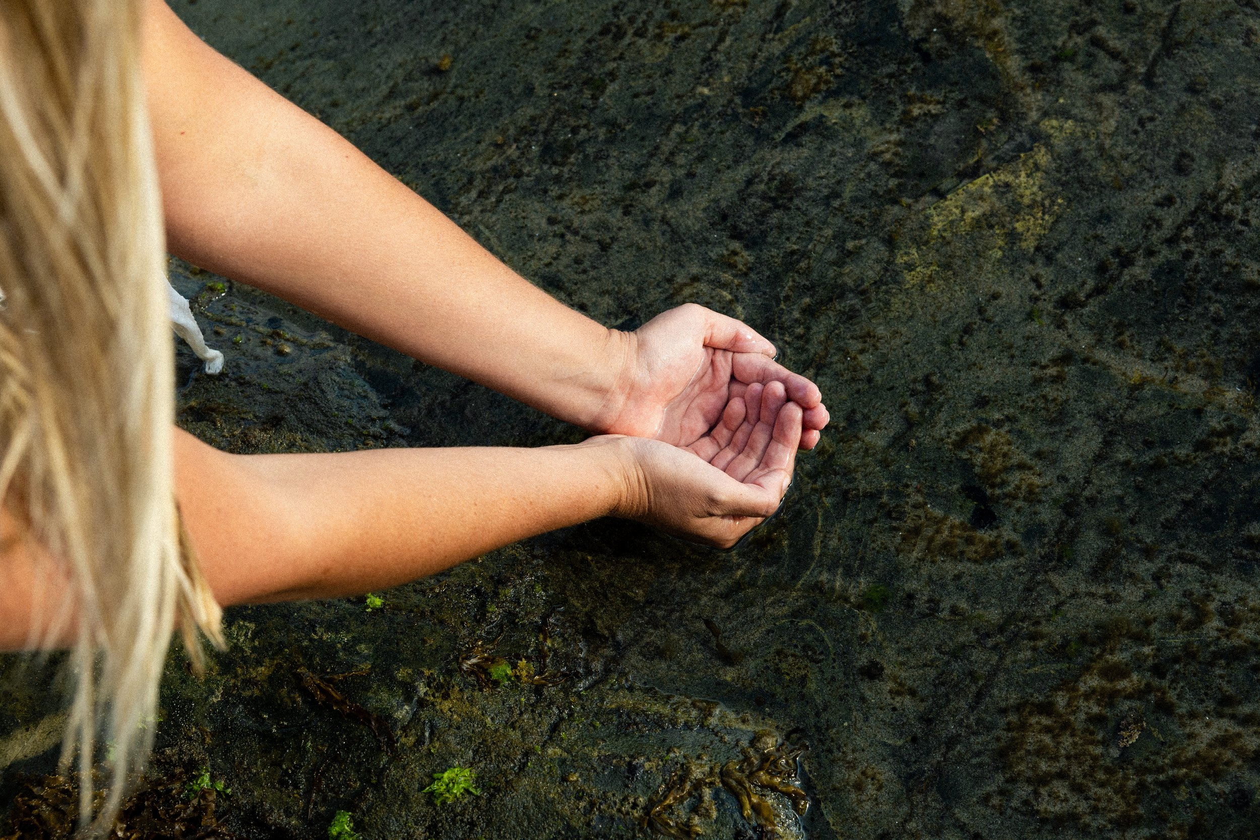 A person with blonde hair is holding their hands together above shallow water, with dark, mossy rocks visible beneath the water surface.