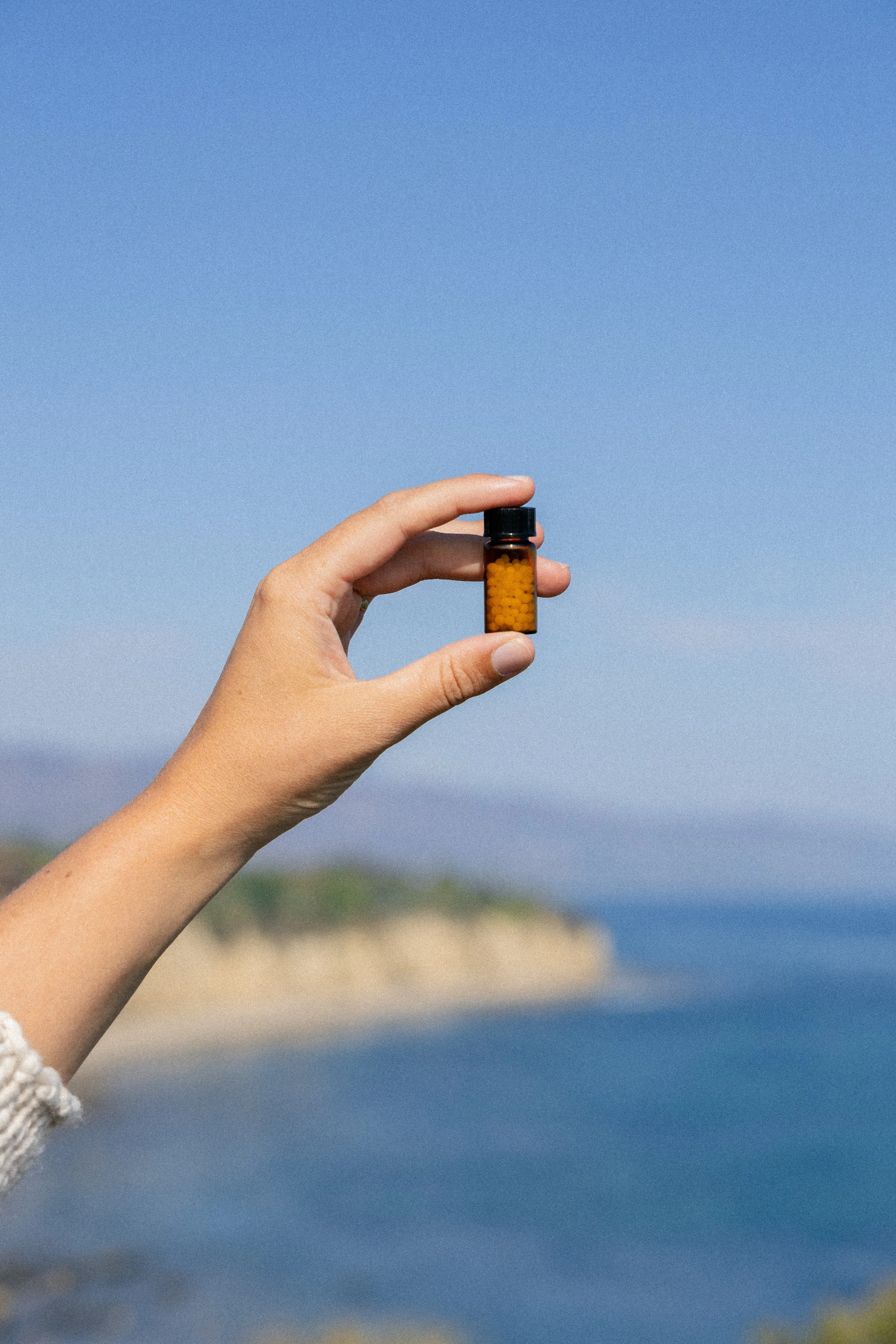 A hand holding a small amber prescription pill bottle with yellow pills against a beach and ocean background.