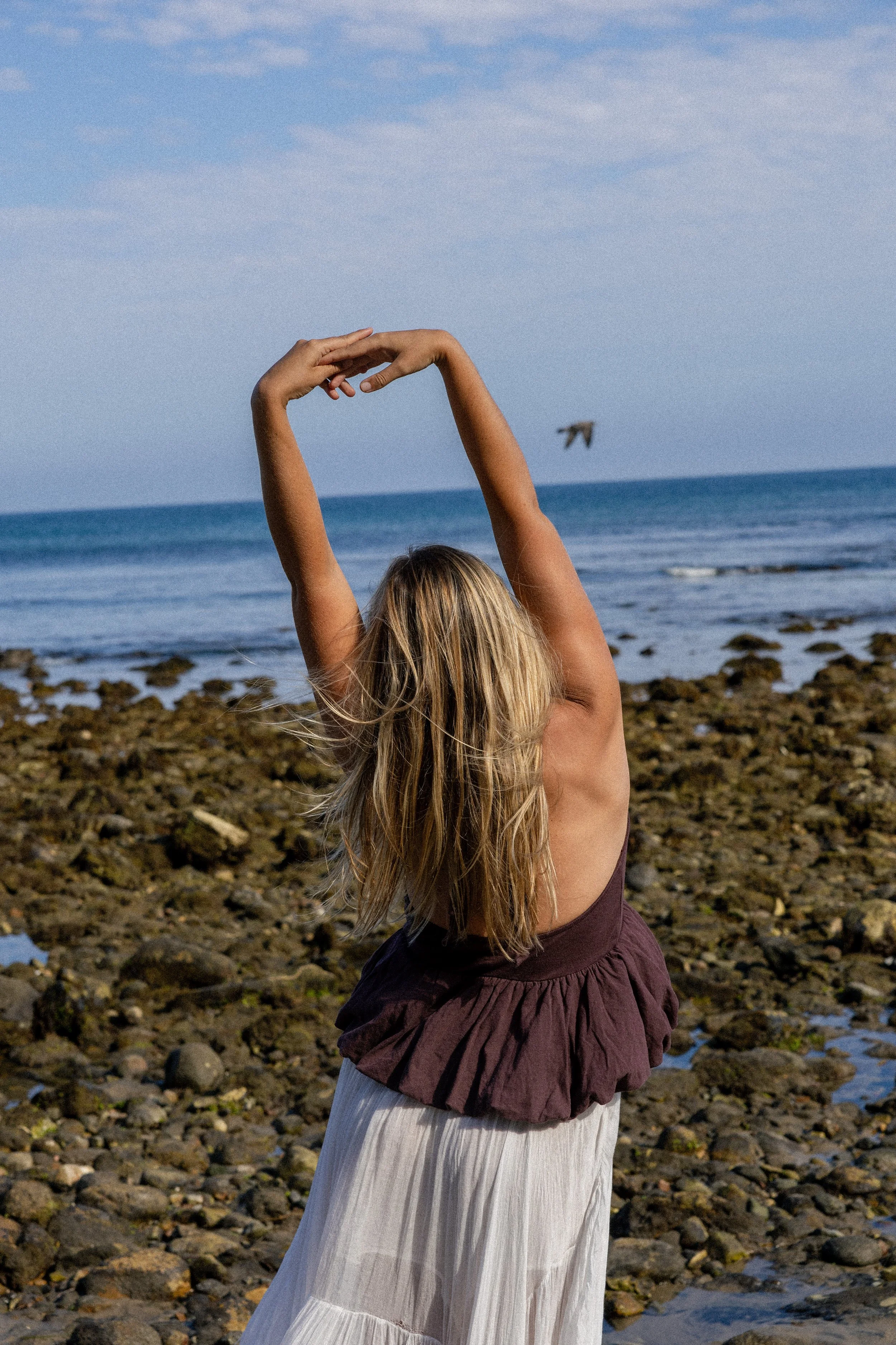 A woman with blonde hair stretching her arms above her head on a rocky beach, with the ocean and a cloudy sky in the background.