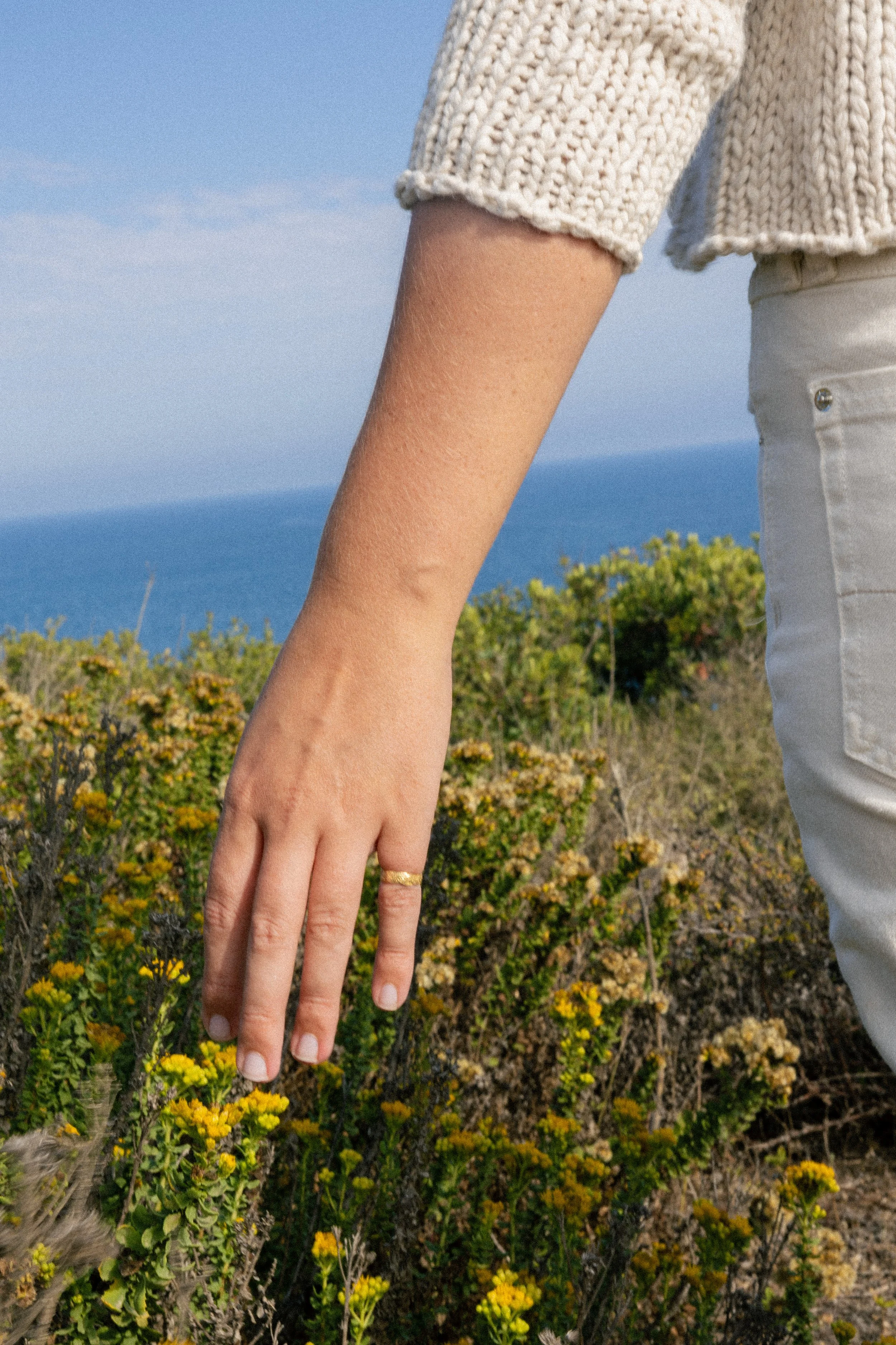 Close-up of a person's hand with a gold ring, hanging down near yellow flowering plants on a coastal background with the ocean and sky.