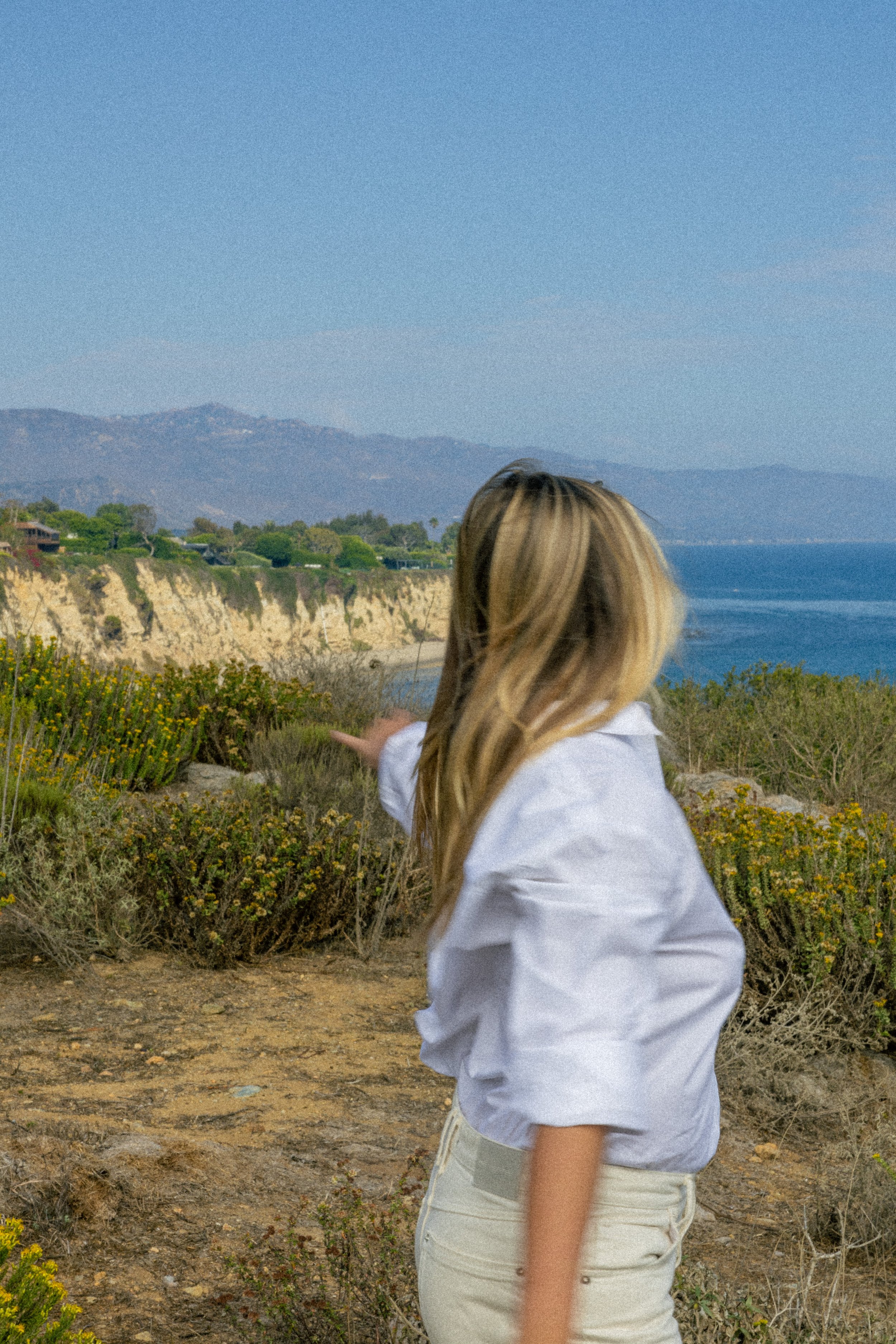 A woman pointing towards the ocean near a cliff with mountains in the background.