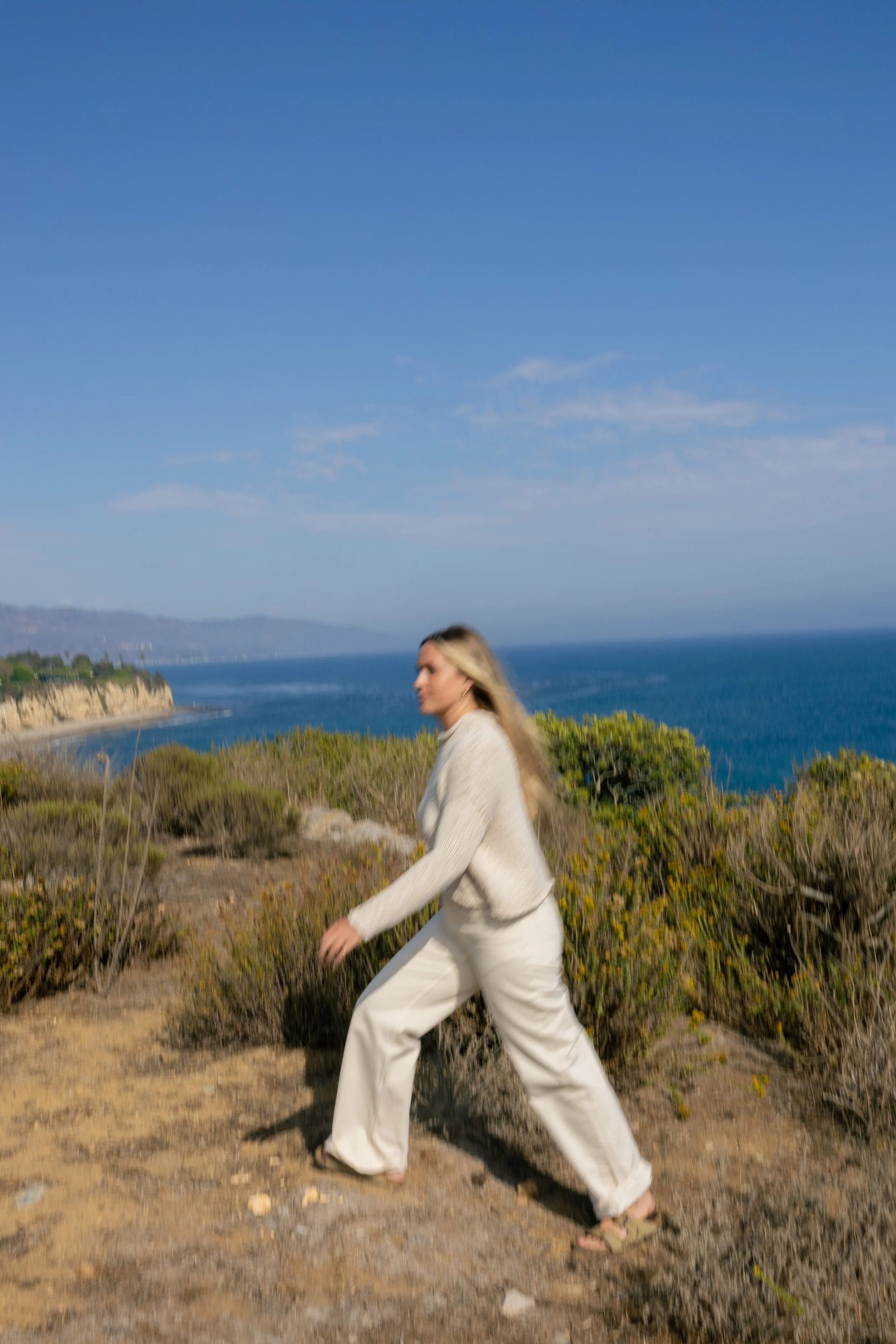 A woman in white pants and a beige sweater walking along a coastal bluff with the ocean and cliffs in the background.