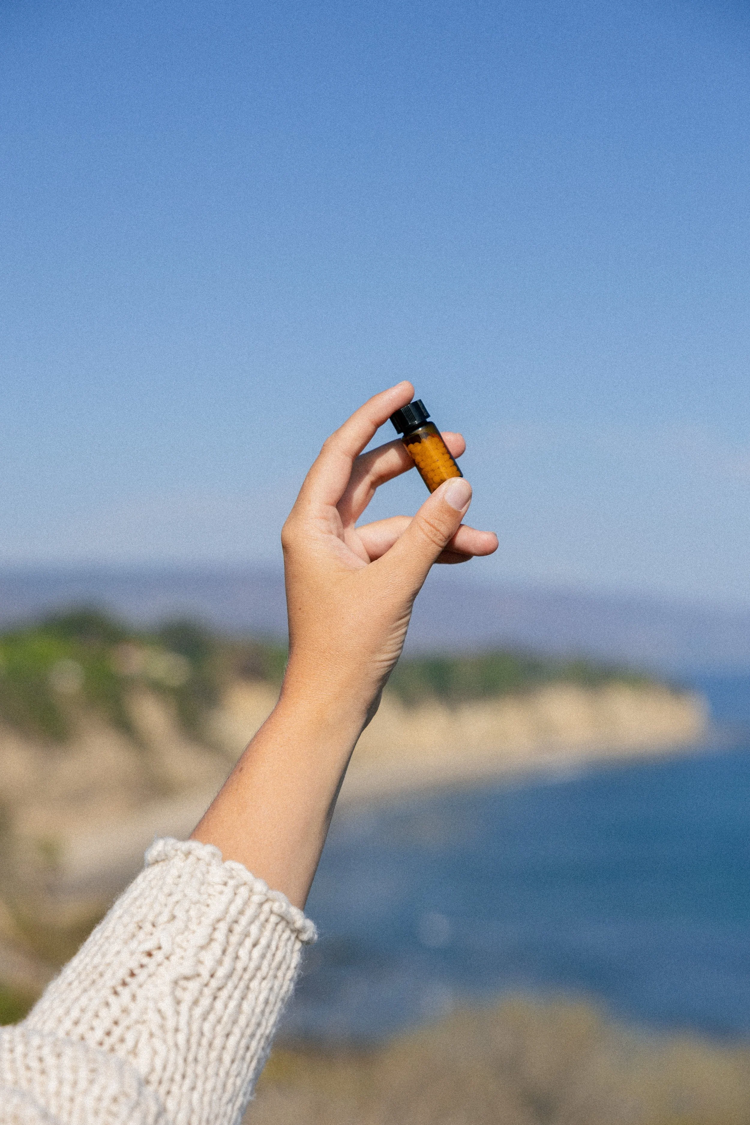 A hand holding a small amber medication bottle with a black cap against a scenic coastal background with sky, cliffs, and ocean.