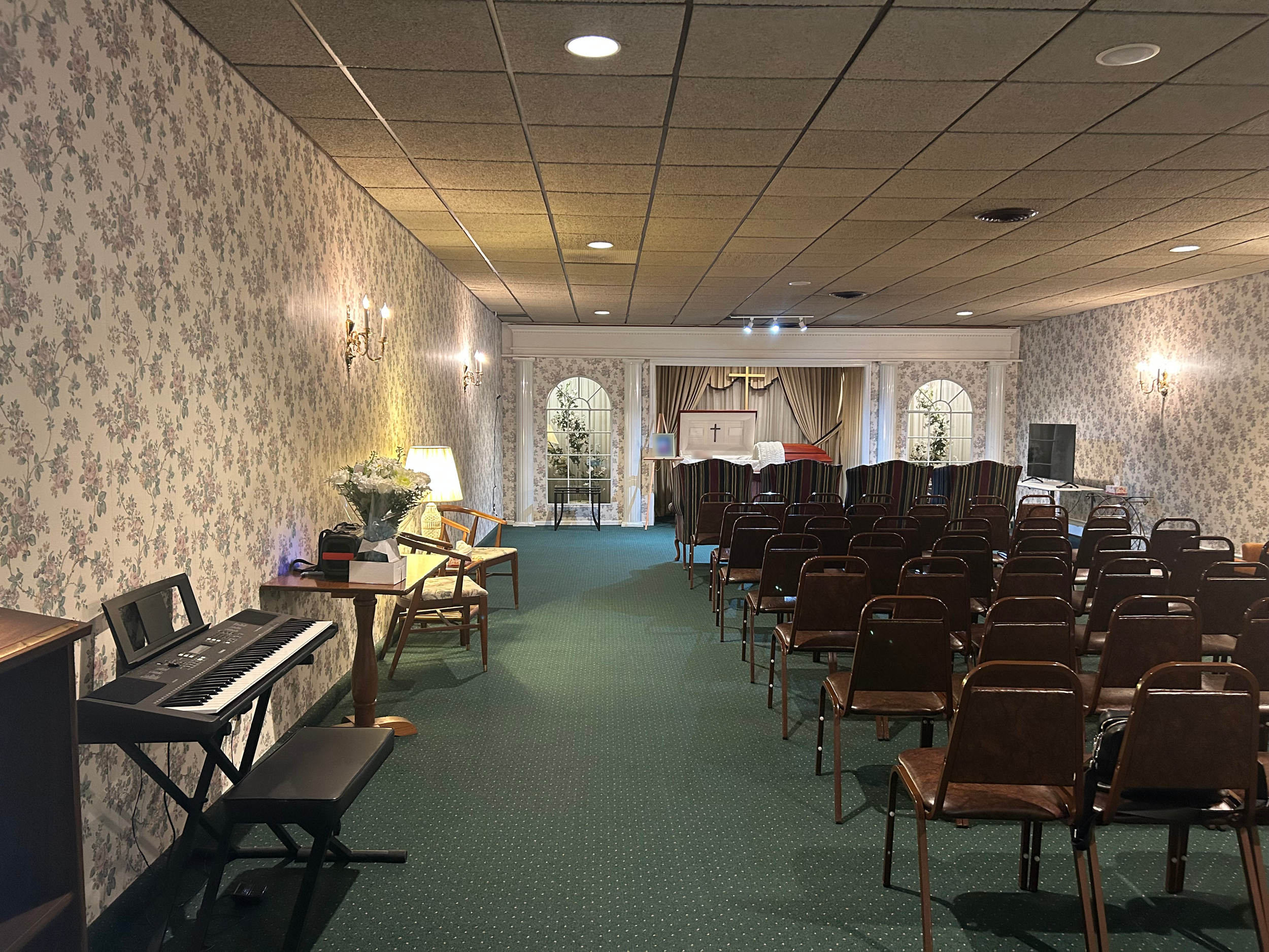 Inside view of a small chapel or prayer room with floral wallpaper, brown chairs arranged facing an altar with a cross, a piano on the left, and a small table with flowers on it.