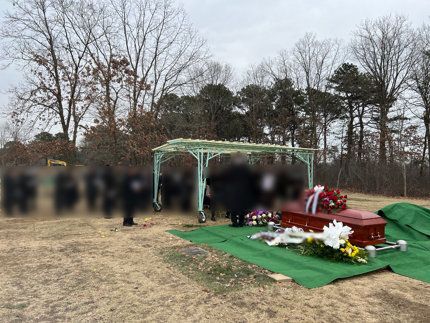 A funeral scene outdoors with a coffin draped in a flag, surrounded by flowers and floral arrangements, under a metal canopy. Several blurred individuals are attending, with a backdrop of trees and overcast sky.