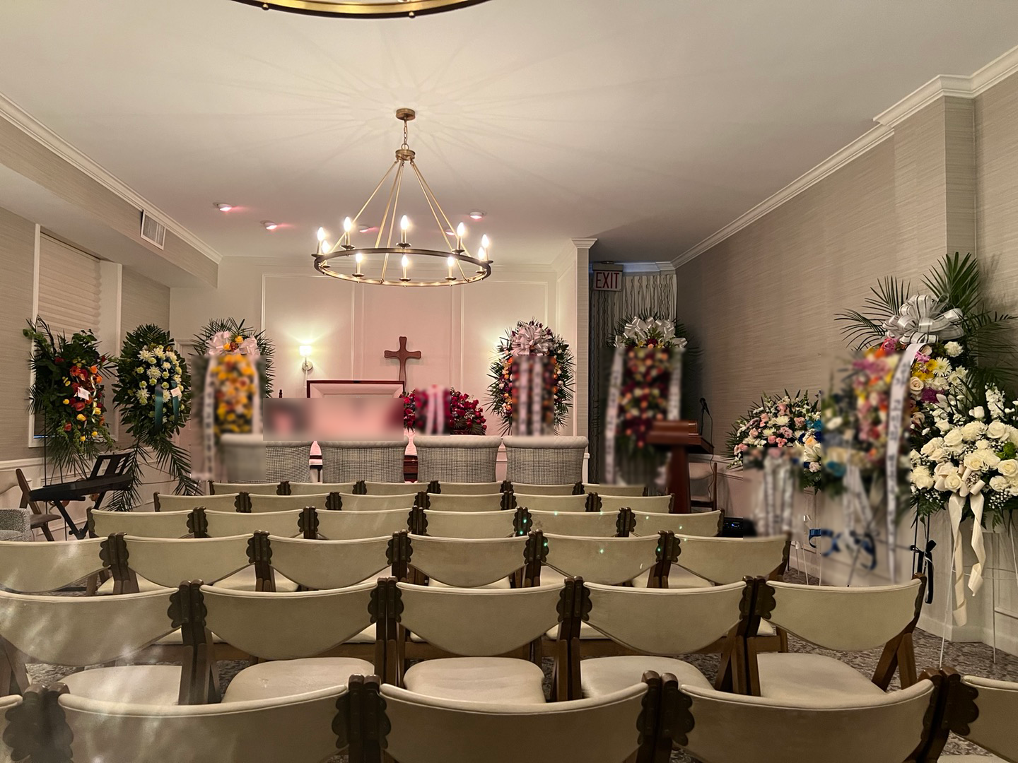 Interior of a funeral home or memorial service room decorated with numerous floral arrangements, a wooden cross at the front, and chairs arranged in rows facing the altar.