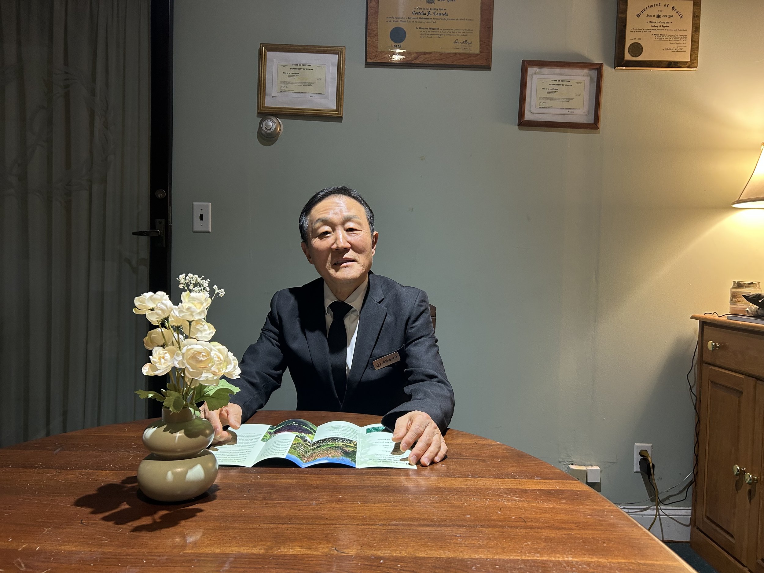 A man in a suit sitting at a wooden table with an open book and a vase of white flowers, in a room with framed certificates on the wall and a lamp on a wooden cabinet.