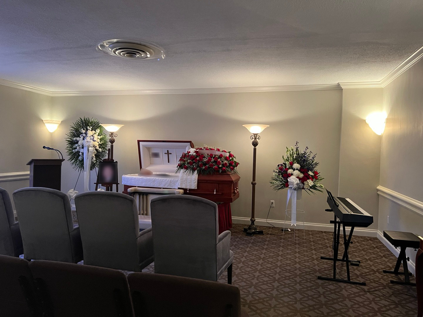 Funeral home setting with an open casket surrounded by flower arrangements, velvet chairs in front, and a piano on the side.
