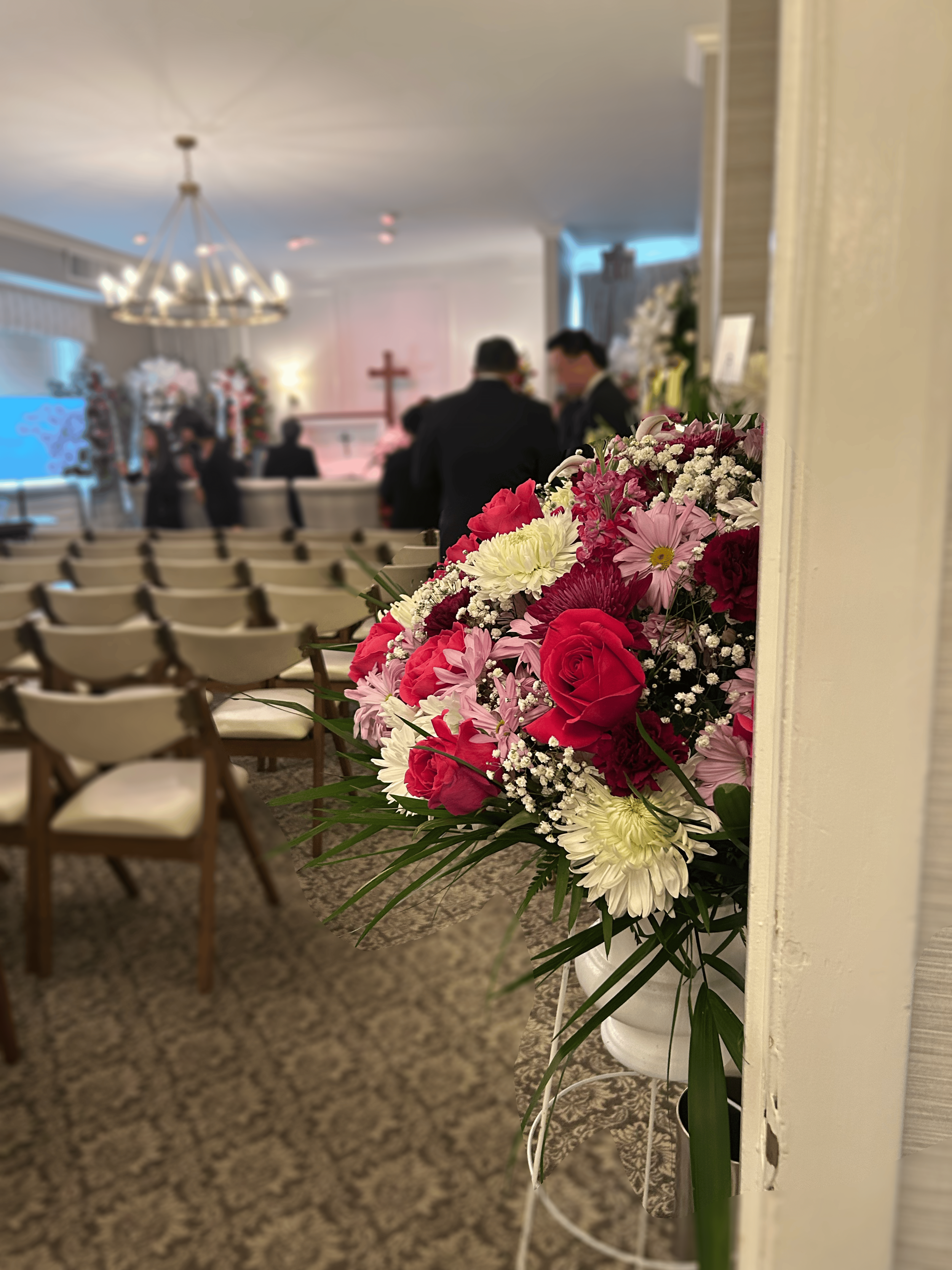 A floral arrangement near a wall in a church or event hall with chairs, people dressed in formal attire, and a cross on the altar in the background.