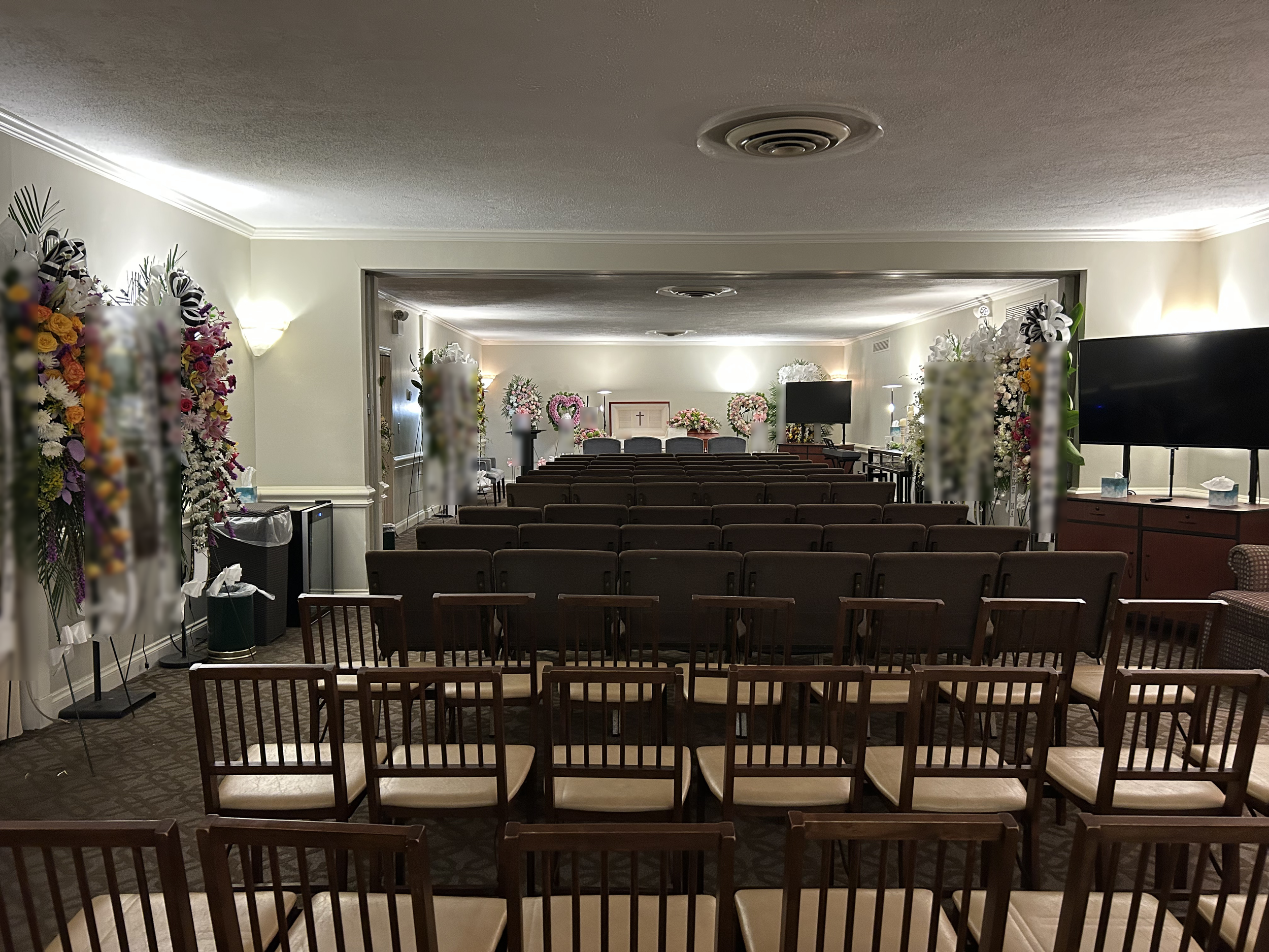 Funeral service room decorated with flower arrangements, chairs arranged facing a casket draped with a cloth, and a cross on the wall at the front.