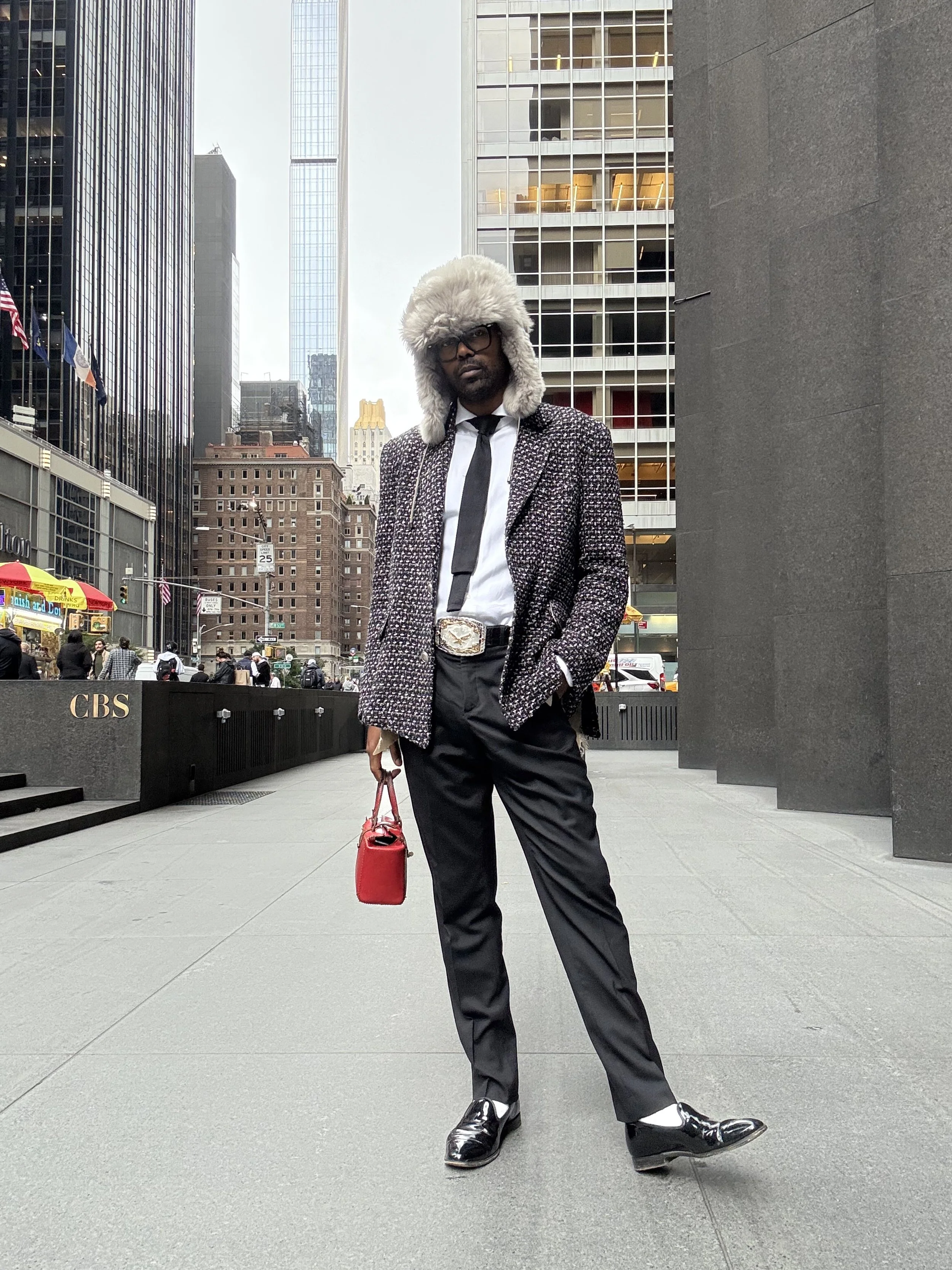 A man in a patterned blazer, white shirt, black tie, and black trousers standing on a city sidewalk. He wears a furry hat, glasses, and shiny black shoes, holding a red handbag in his right hand. Tall buildings and a CBS sign are visible in the backg