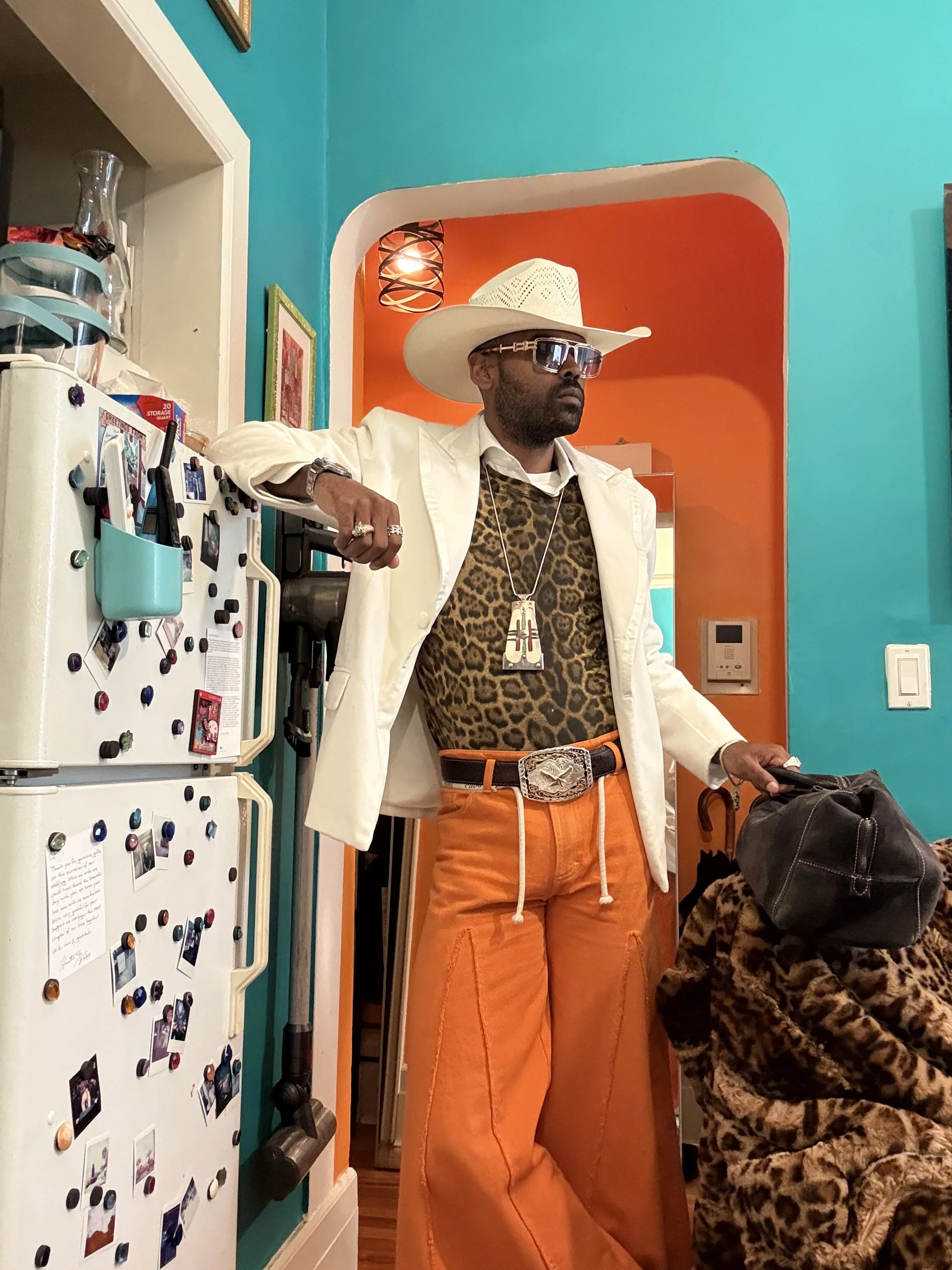Man wearing a wide-brimmed hat, sunglasses, a white blazer, leopard print shirt, orange pants, and a large belt with a decorative buckle, standing in a brightly colored room.