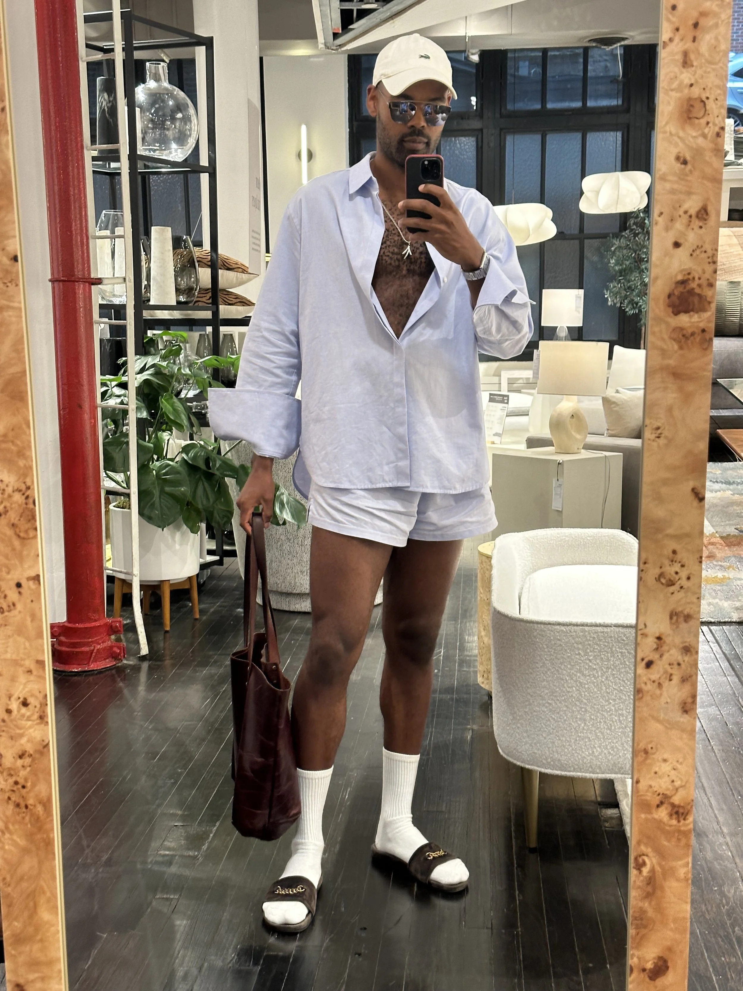 Man taking a mirror selfie in a home decor store, dressed in a white oversized shirt, white shorts, white socks, and slides, wearing sunglasses, a white cap, and carrying a brown tote bag.